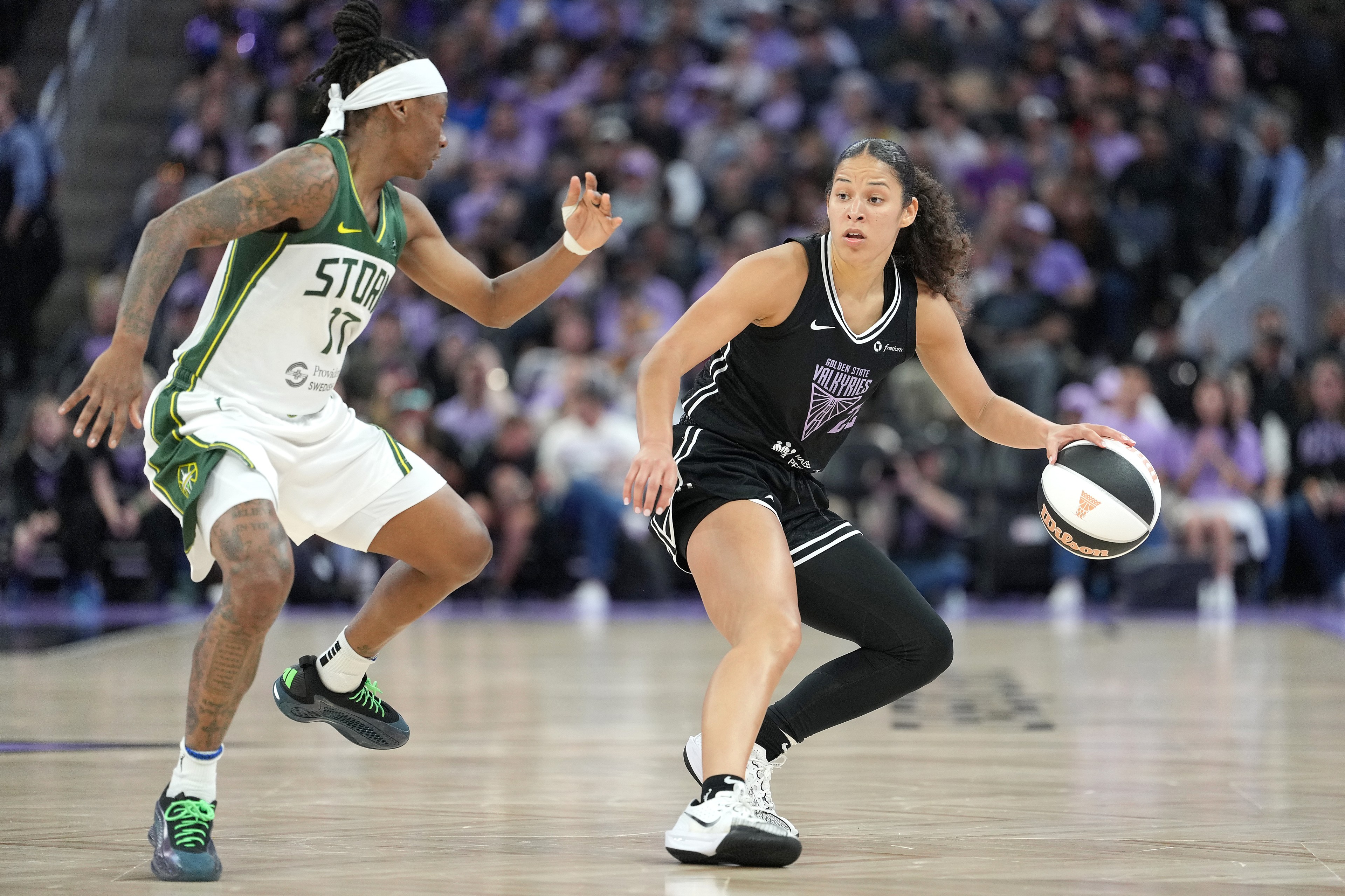 Two basketball players are on a court. One is in a green and white uniform, defending the other, who is dribbling the ball in a black uniform.