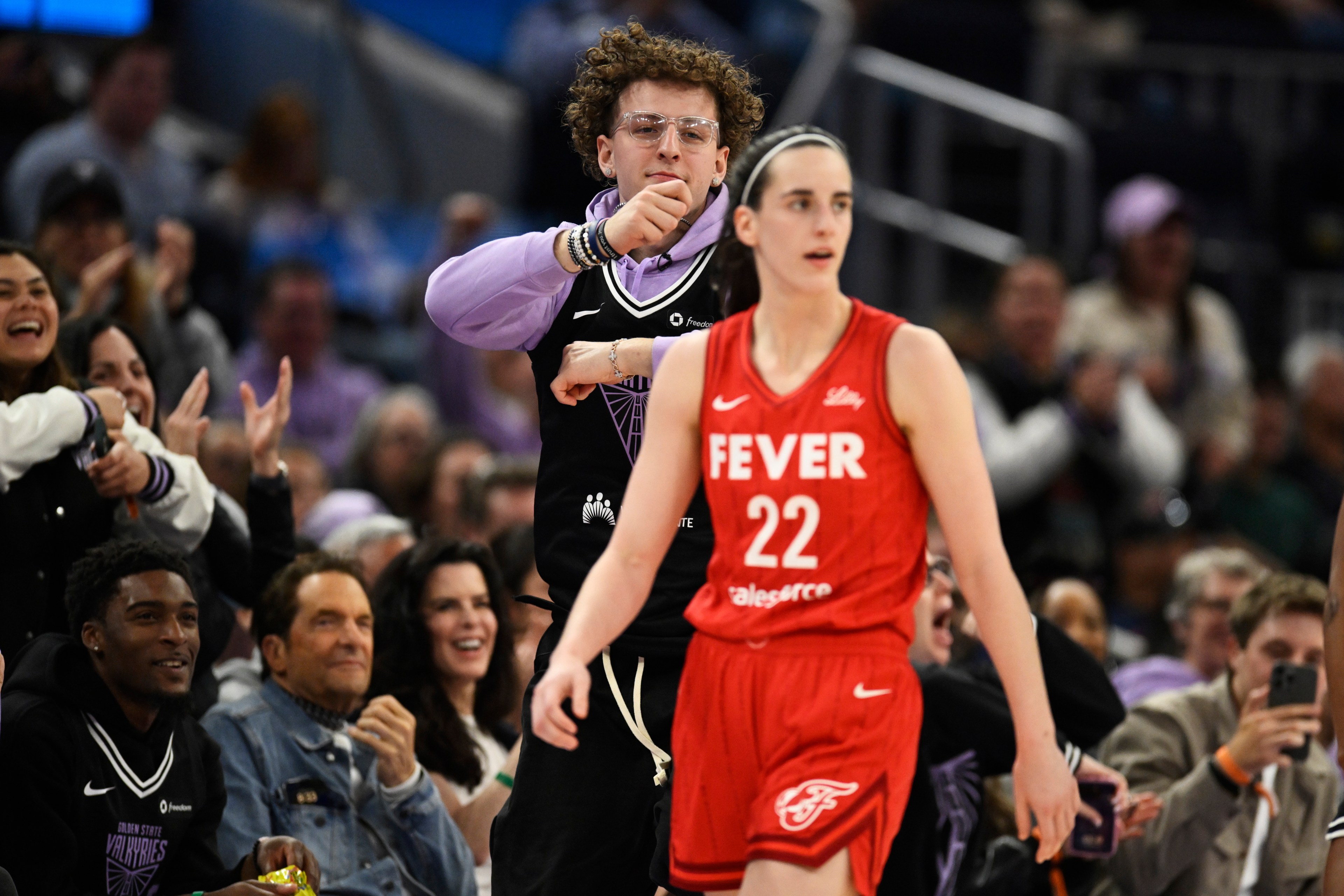 A basketball player in a red uniform walks on the court, while a cheering crowd, including a person in a purple and black jersey, shows excitement in the background.