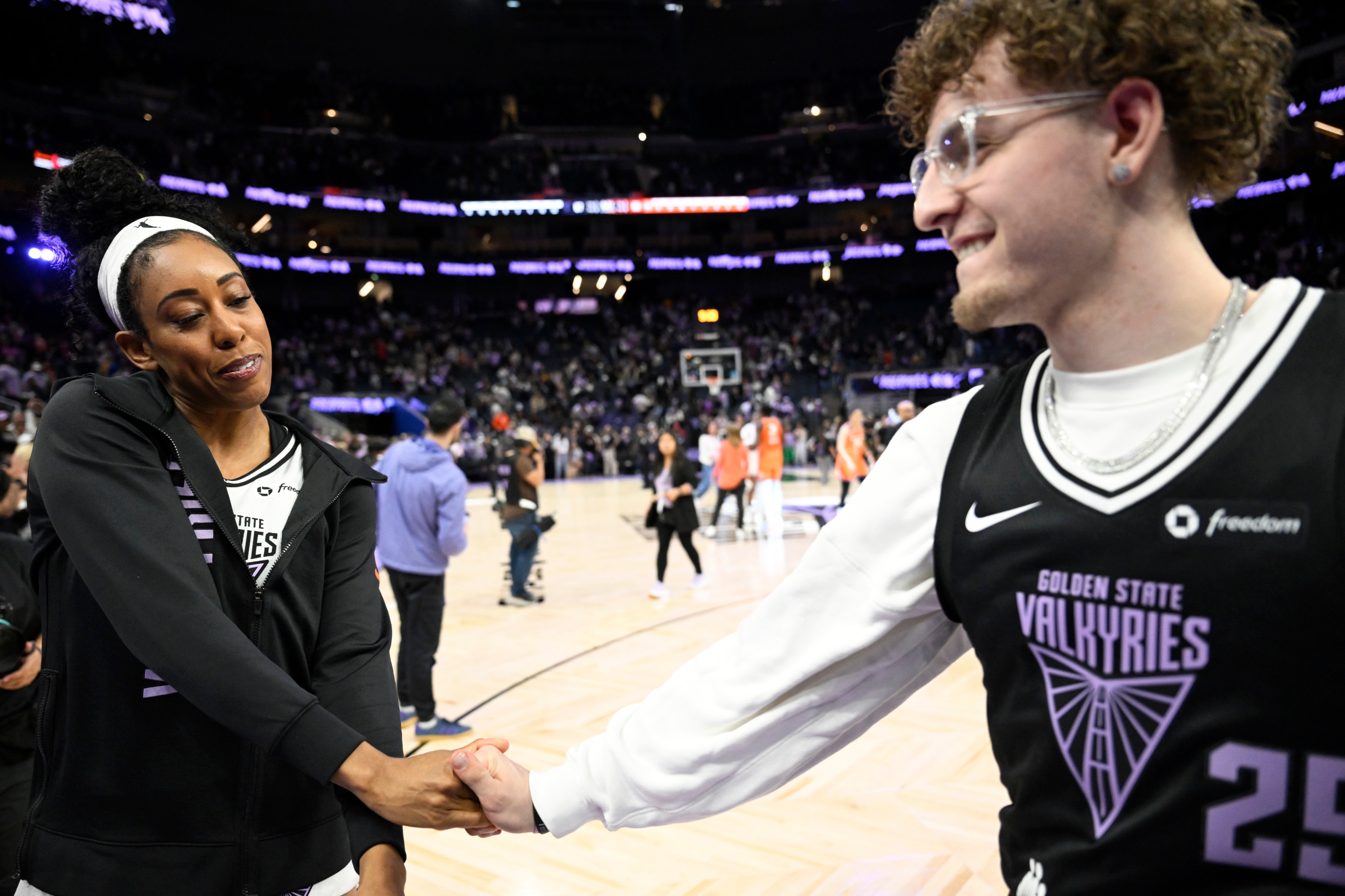 Two people are on a basketball court shaking hands, with a crowd in the background. One wears a &quot;Golden State Valkyries&quot; jersey and the other a black jacket.