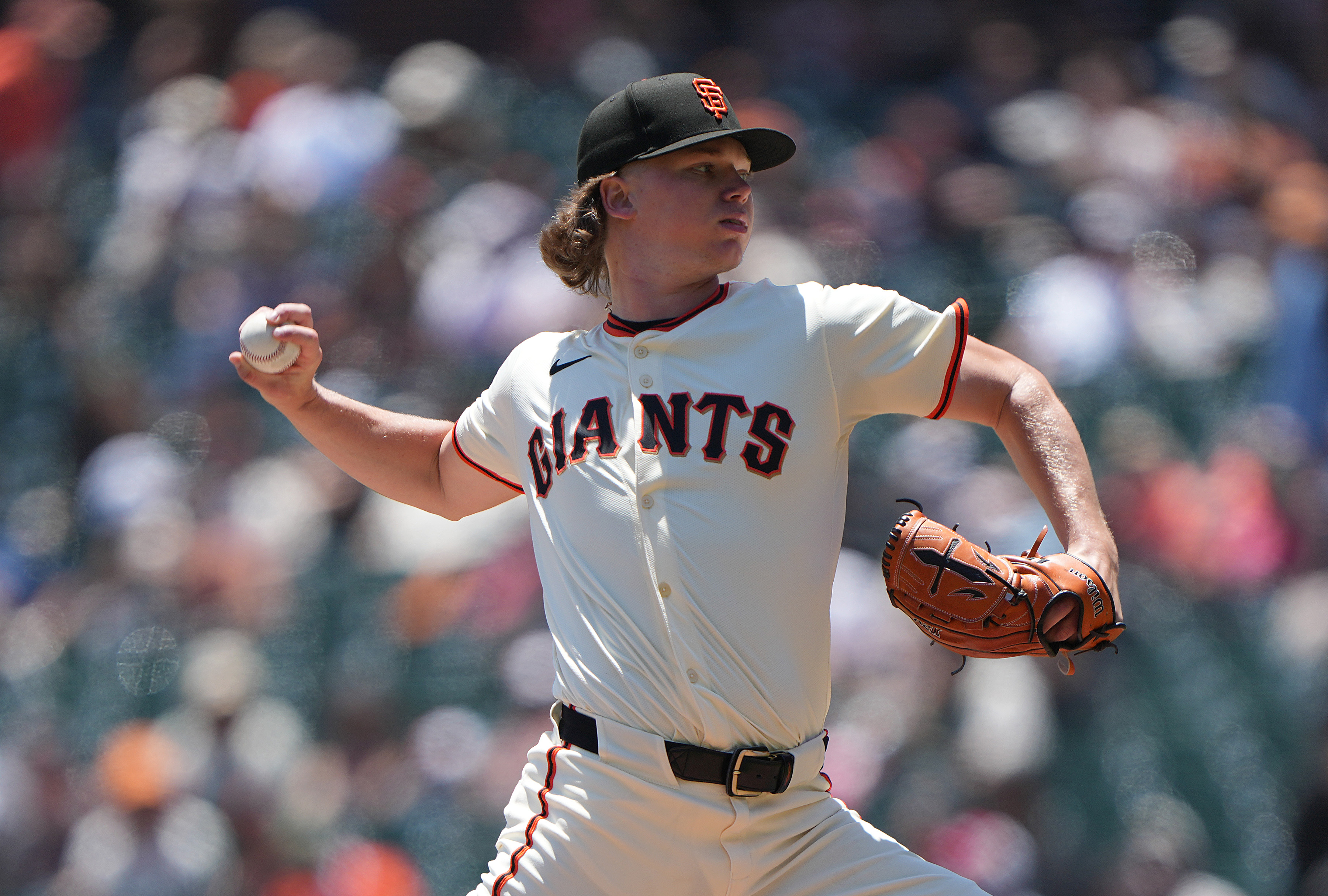 A baseball player in a "Giants" uniform is pitching on a sunny day, holding a ball in one hand and wearing a glove on the other, with a crowd blurred in the background.