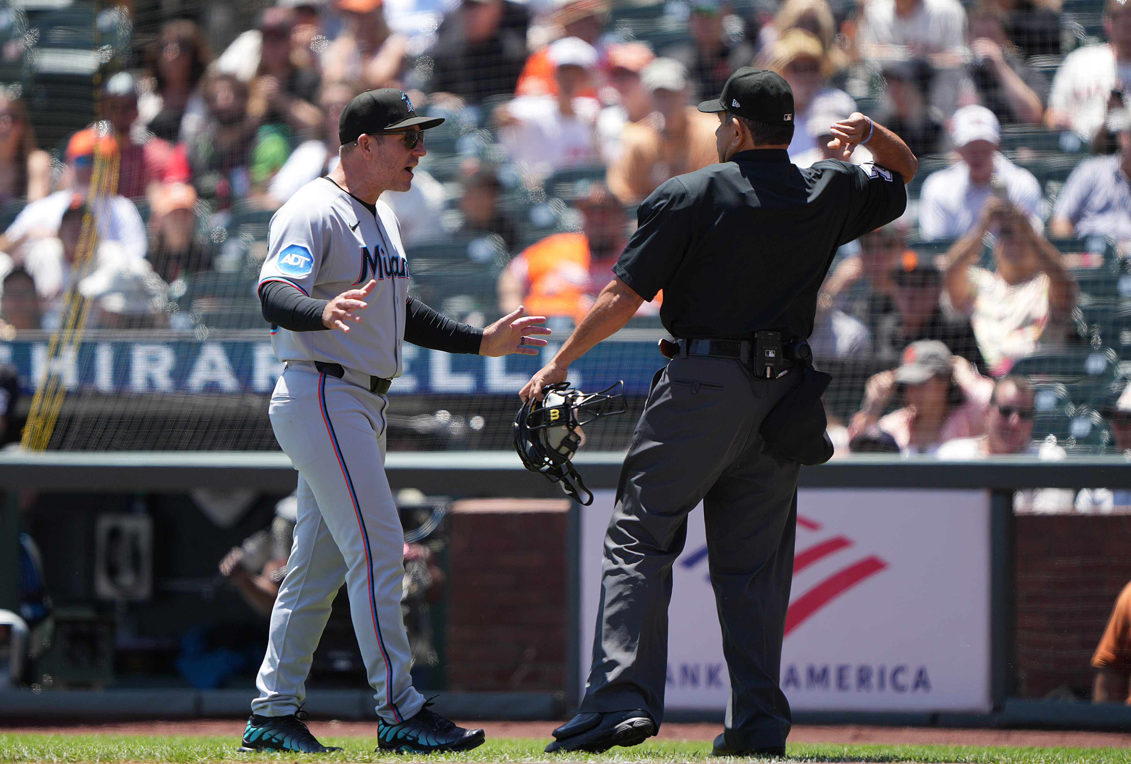 A baseball coach, wearing a Miami jersey and cap, is animatedly discussing something with an umpire who is holding a mask. They stand on a grassy field.