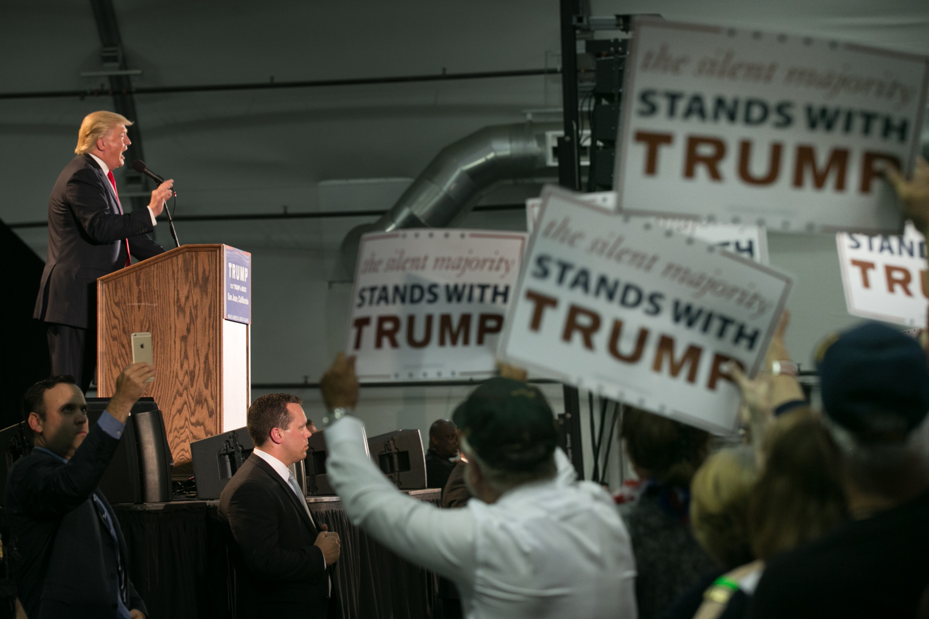 A man at a podium speaks to a crowd holding signs reading, &quot;The silent majority stands with Trump&quot; in a large indoor venue.