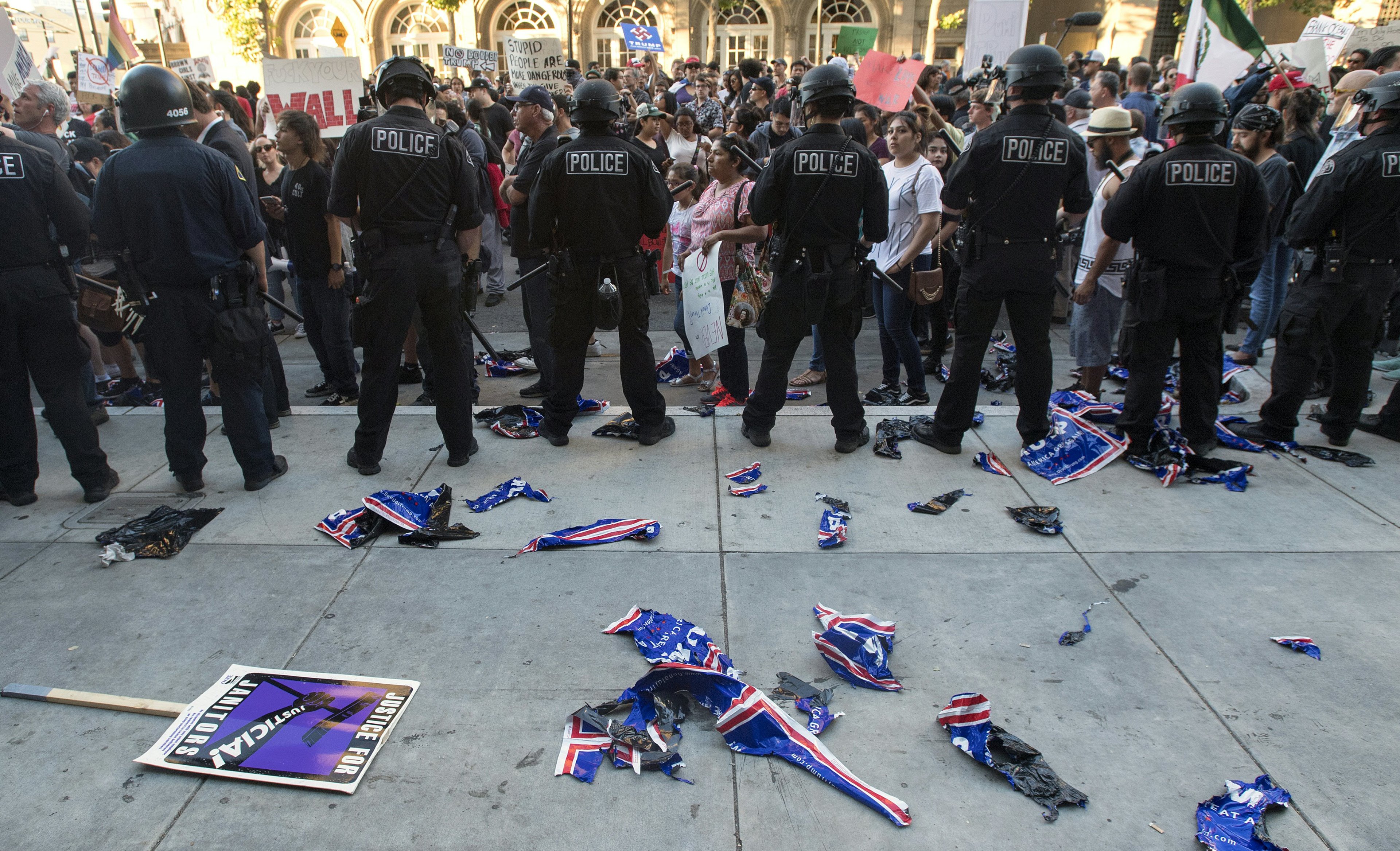 Police officers in riot gear stand in a line facing a crowd of protesters with signs. Torn flags and protest materials litter the ground between them.