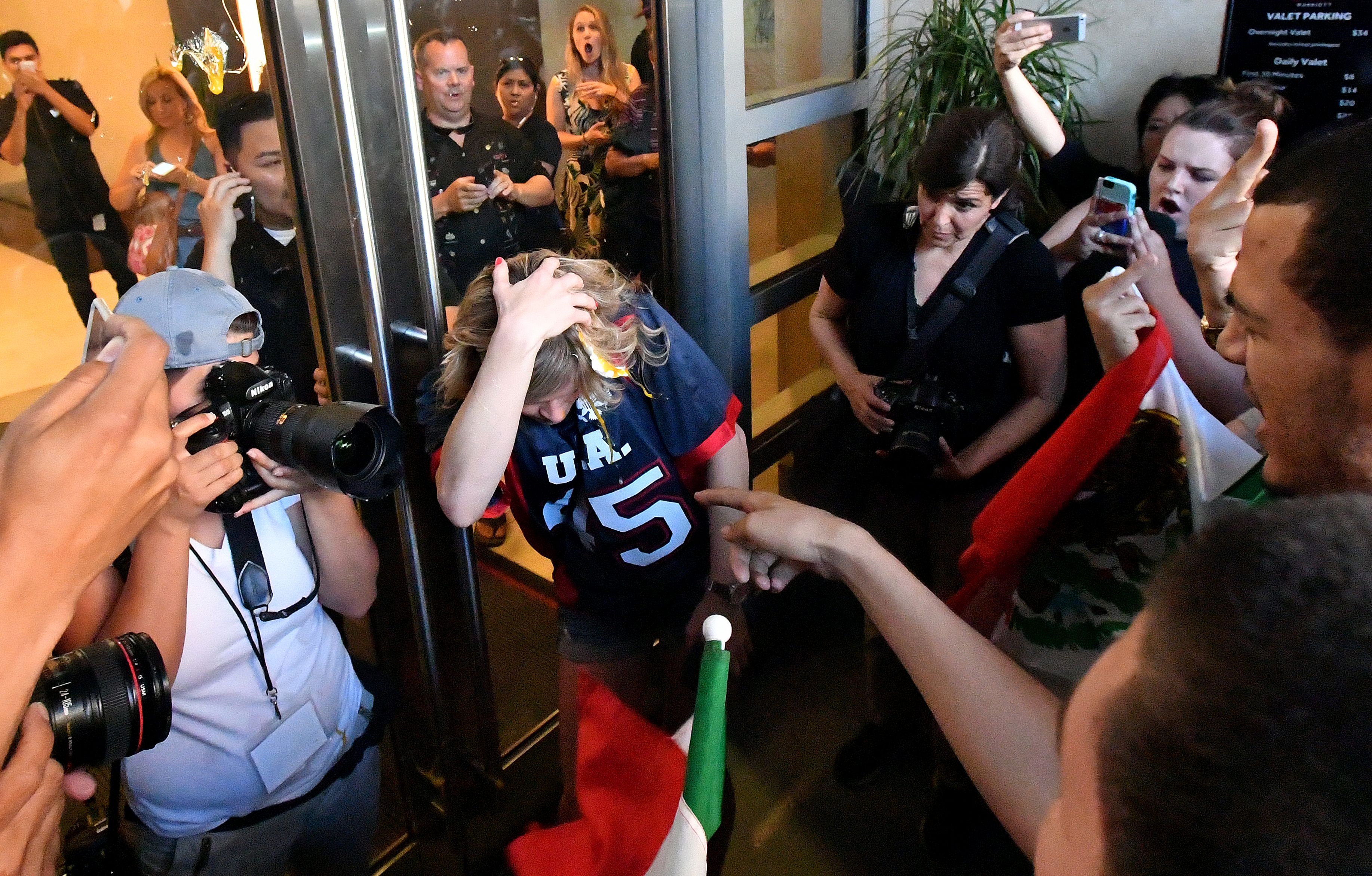 A crowded scene shows a person wearing a &quot;USA 45&quot; jersey being photographed and surrounded by a group, with some holding devices and a flag visible.