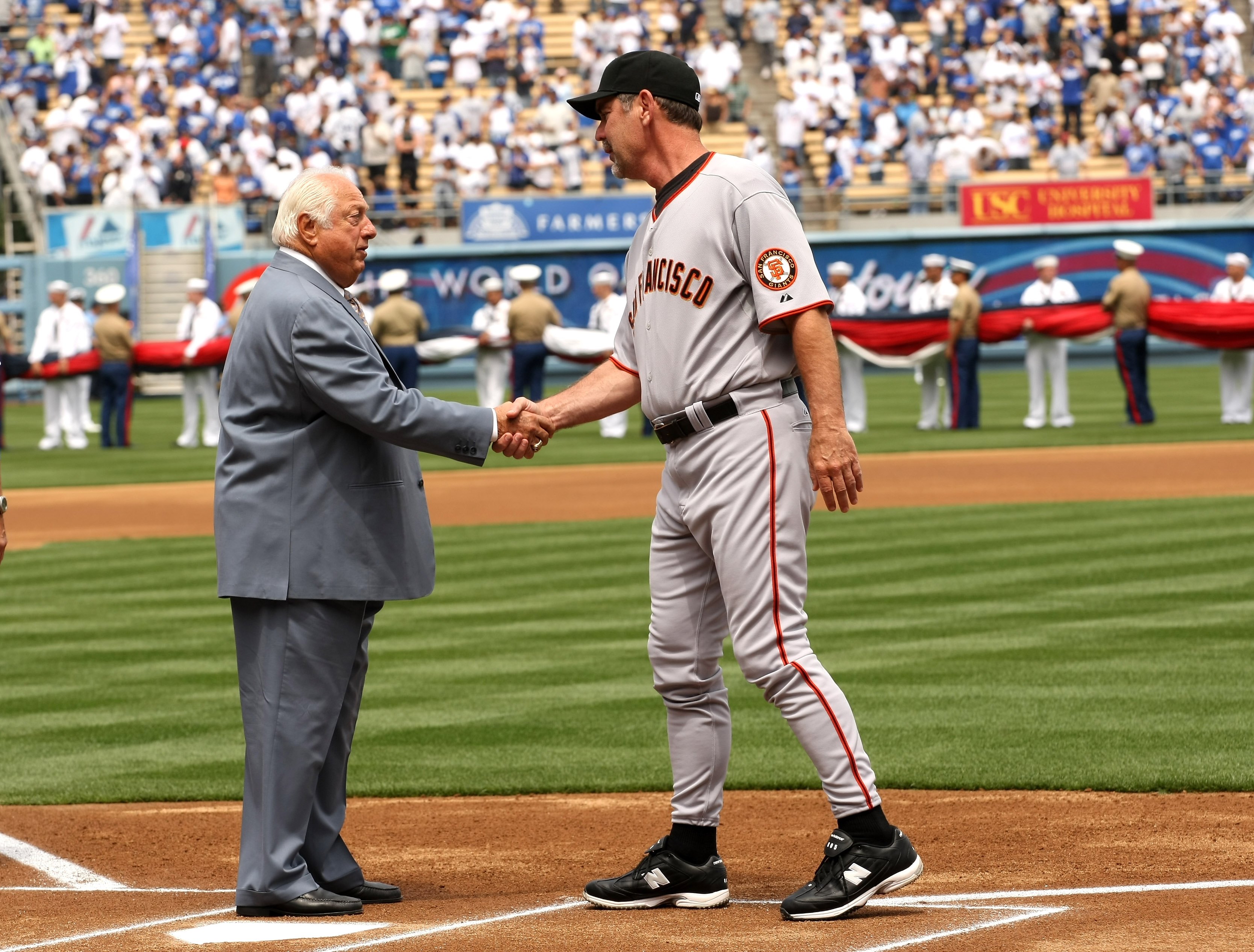 Two men shake hands on a baseball field; one in a gray suit, the other in a gray San Francisco uniform. A flag ceremony and crowd are in the background.