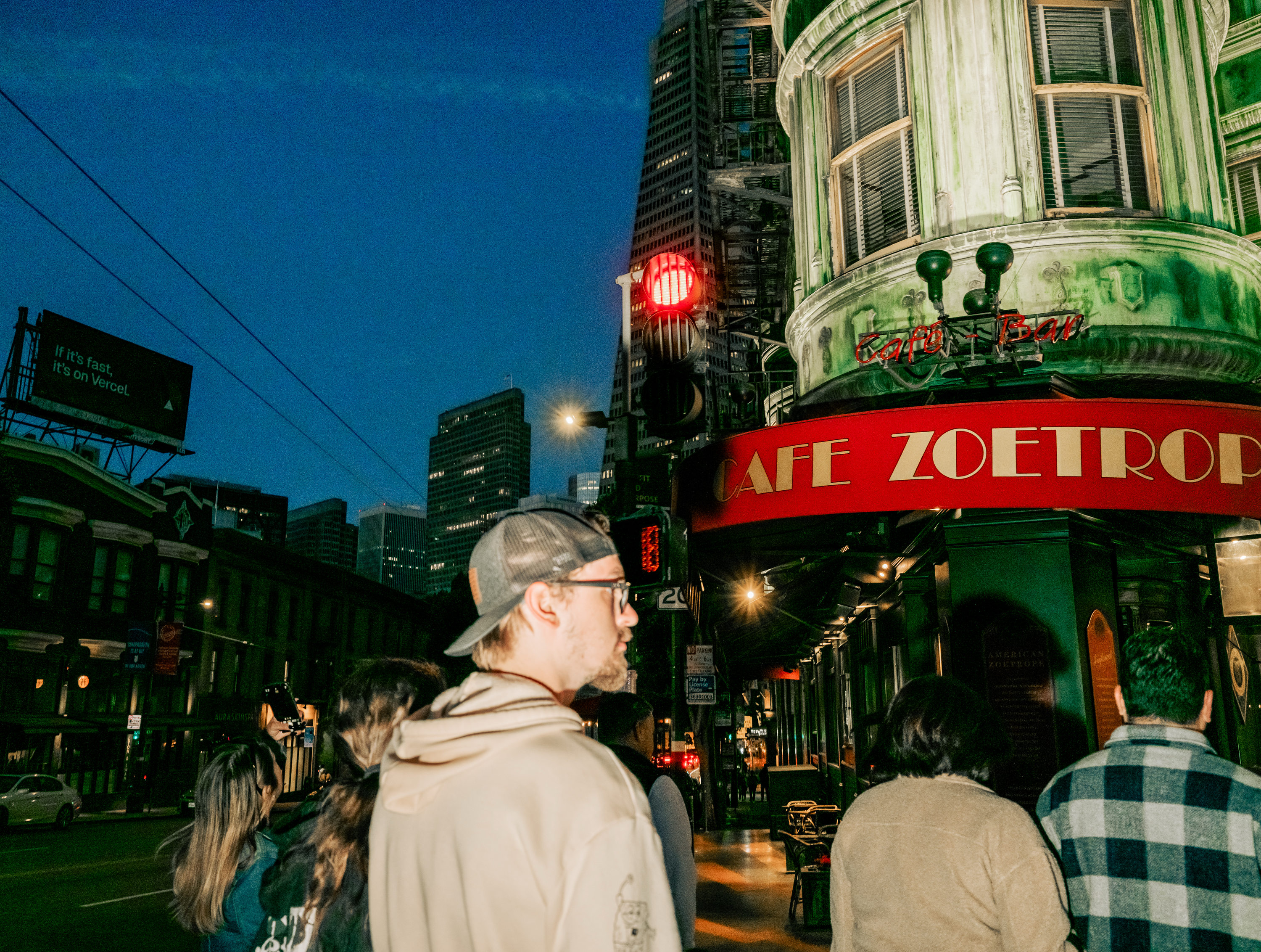 A group of people stand on a city street at night near a building with a red &quot;Cafe Zoetrope&quot; sign. Skyscrapers are in the background under a dark blue sky.
