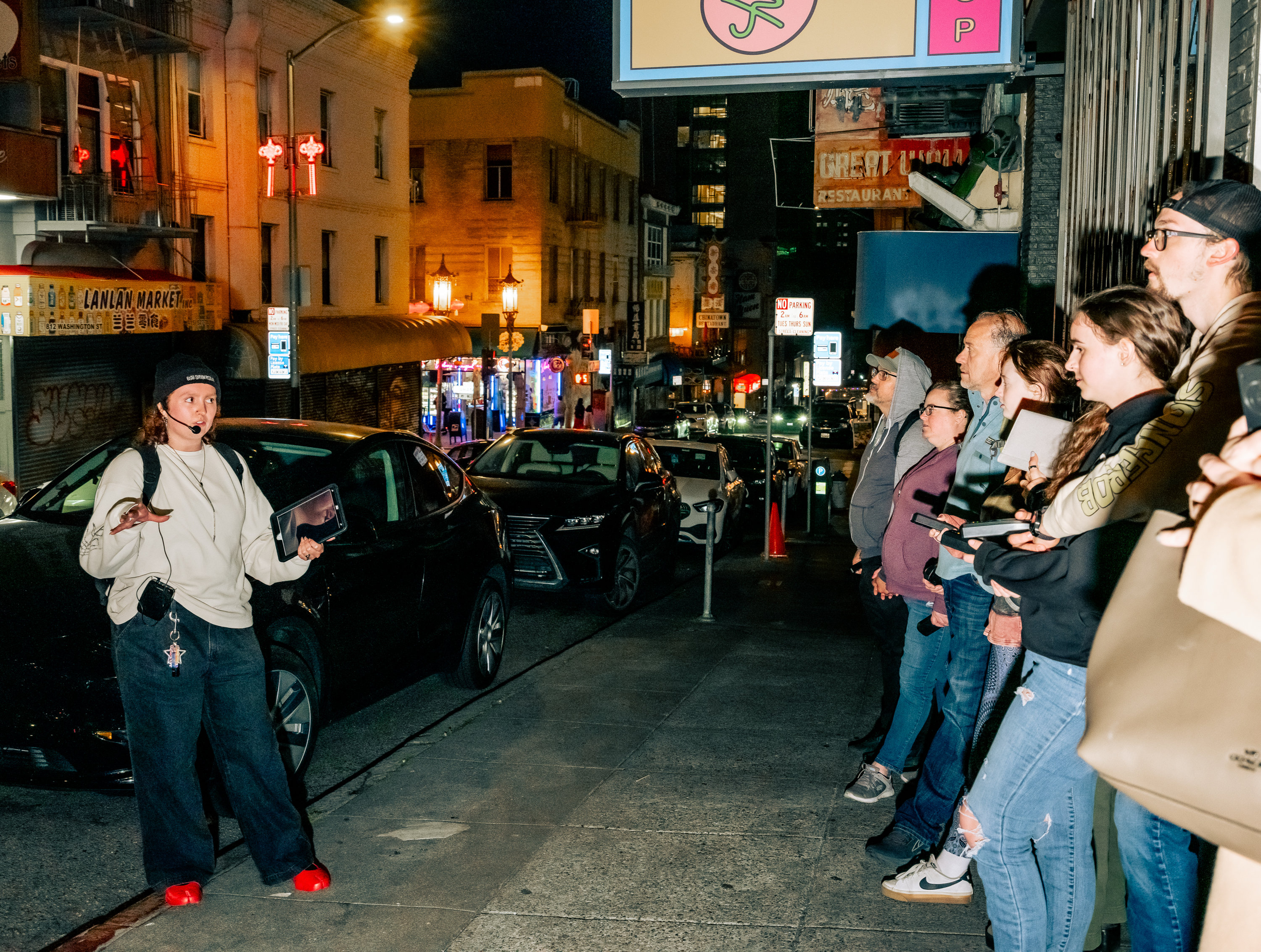 A person in a cap and mic addresses an attentive group on a nighttime street, with parked cars and lit storefronts in the background.