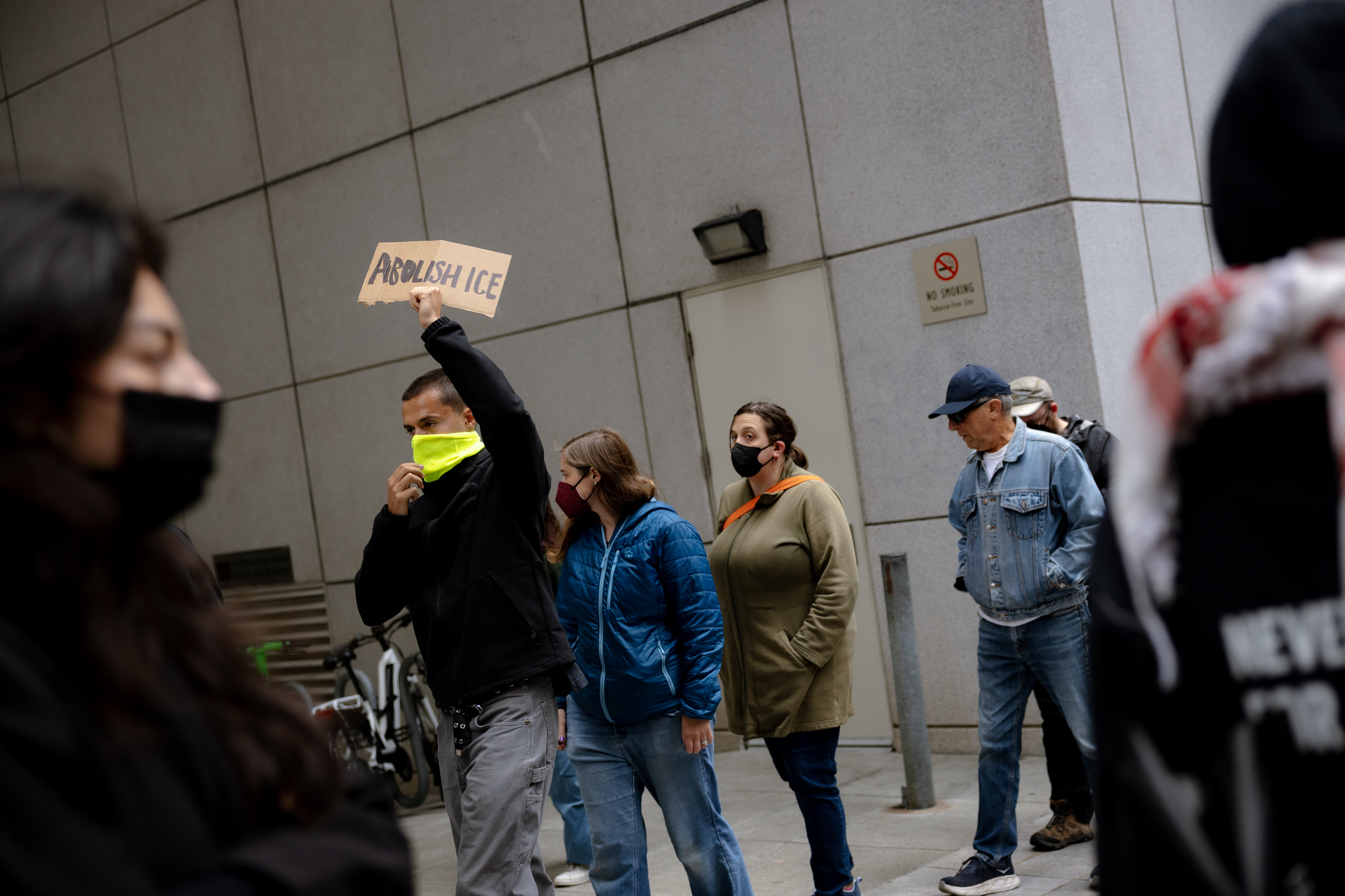 A group of masked individuals stands outside a building, with one person holding a sign reading &quot;Abolish ICE.&quot; They appear to be part of a demonstration.