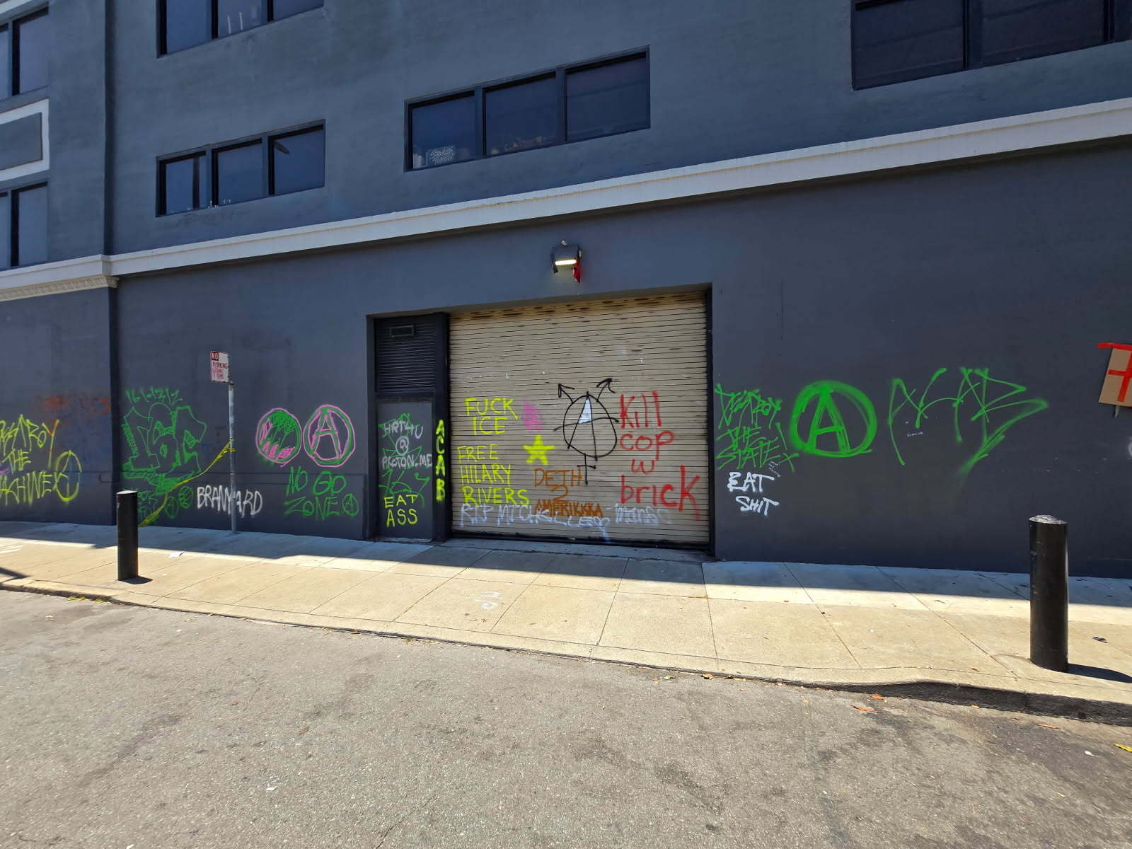 A gray building wall featuring various graffiti with colorful messages and symbols, including anarchist signs and phrases, covers the entire urban scene.