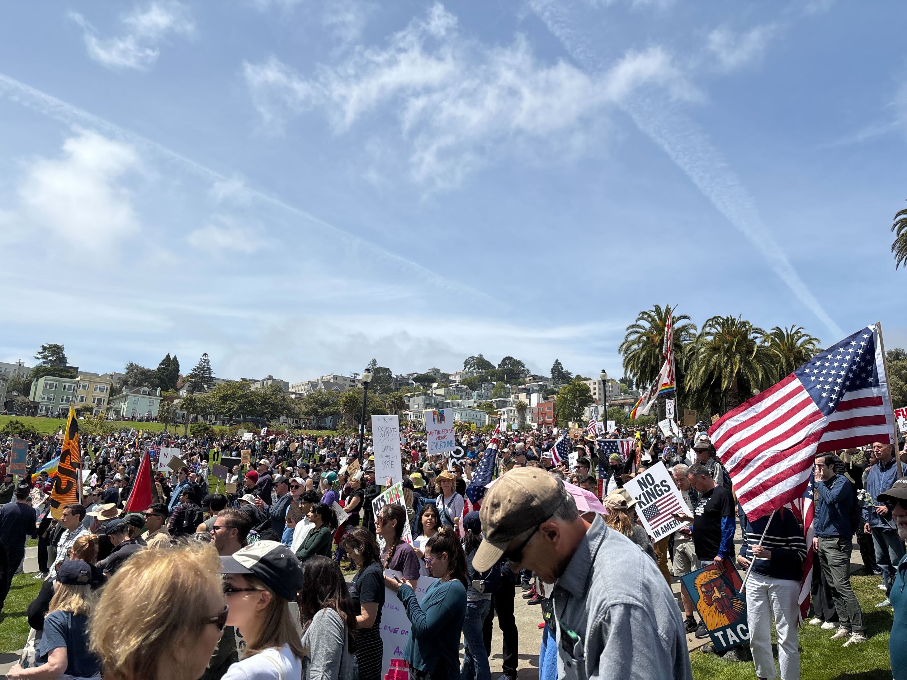 A large crowd gathers in a park under a blue sky, holding signs and flags, including the U.S. flag. Palm trees and buildings are visible in the background.