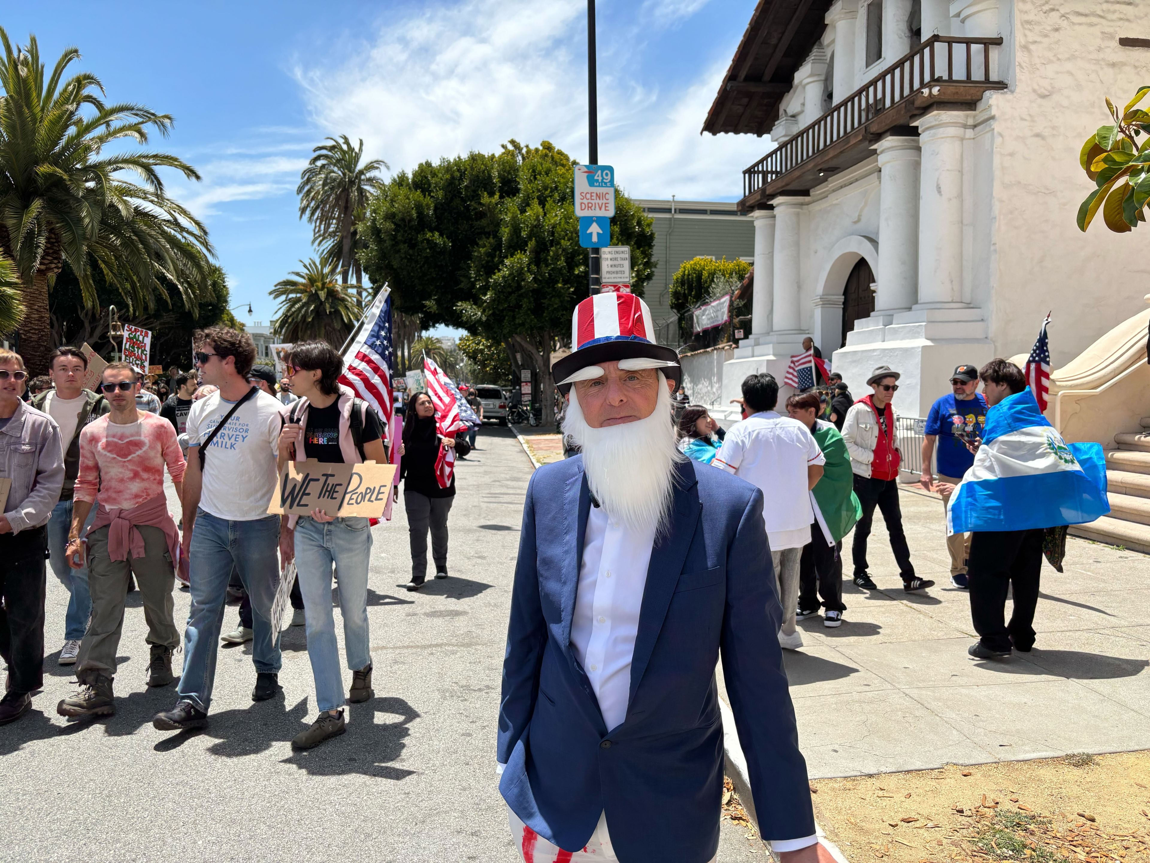A man dressed as Uncle Sam stands in the foreground, while behind him, a group of people with American flags march. One person holds a &quot;We The People&quot; sign.
