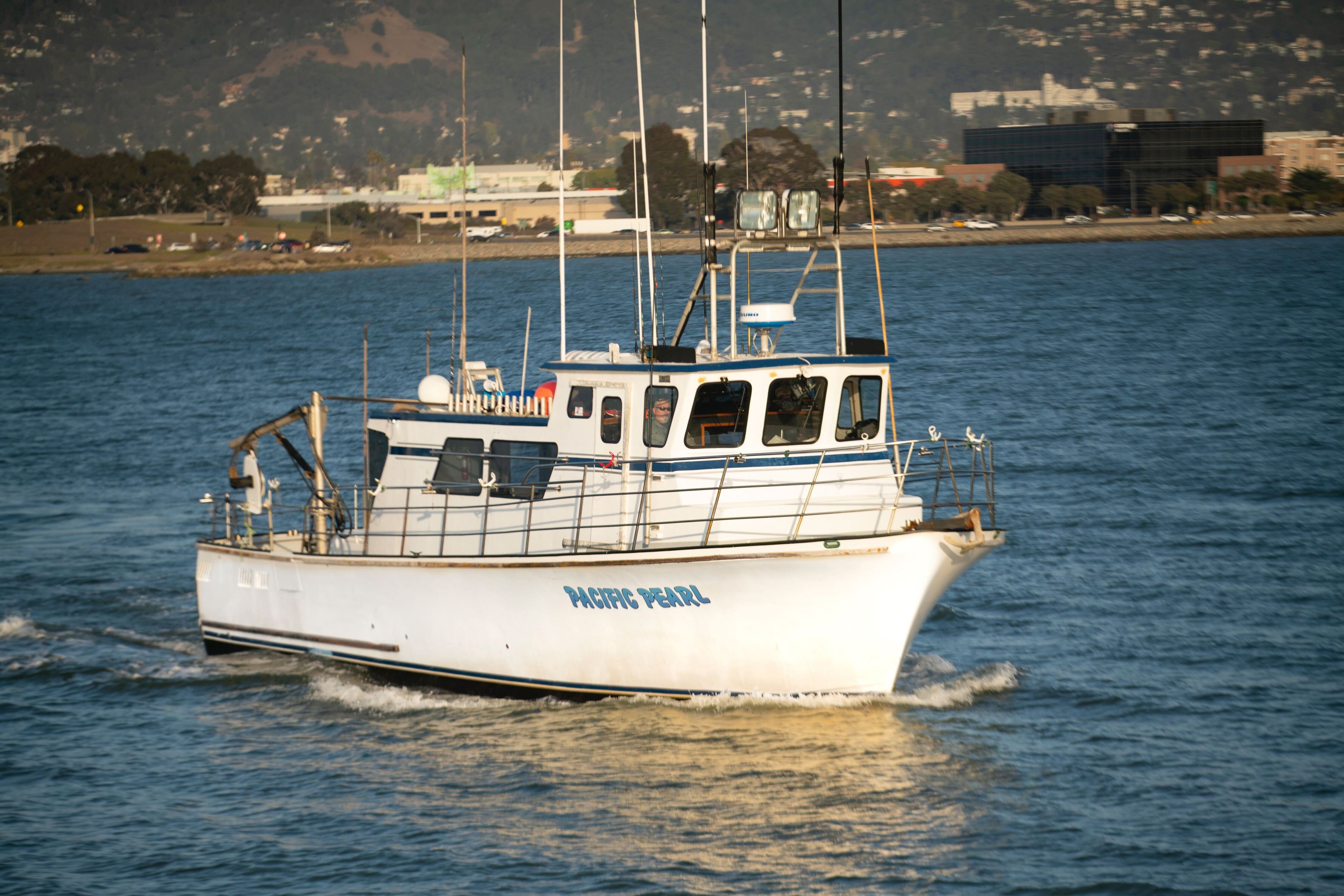A white fishing boat labeled &quot;Pacific Pearl&quot; sails on calm water. Its deck has antennas, and there's a shoreline and buildings in the distant background.