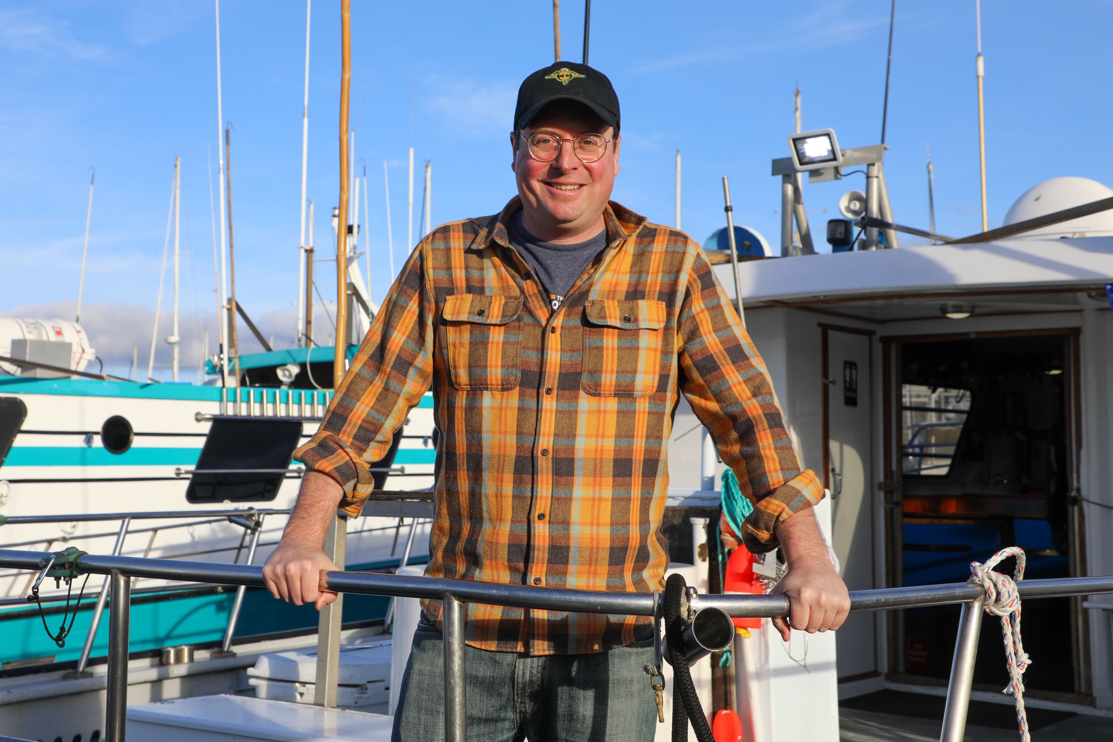 A person in a plaid shirt and cap smiles while standing on a boat, holding onto a railing. The background shows other boats and a clear blue sky.