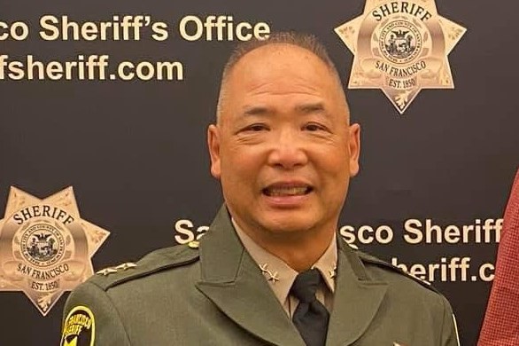 A uniformed officer smiles, standing in front of a San Francisco Sheriff's Office backdrop. The uniform is dark with badges and a name tag.