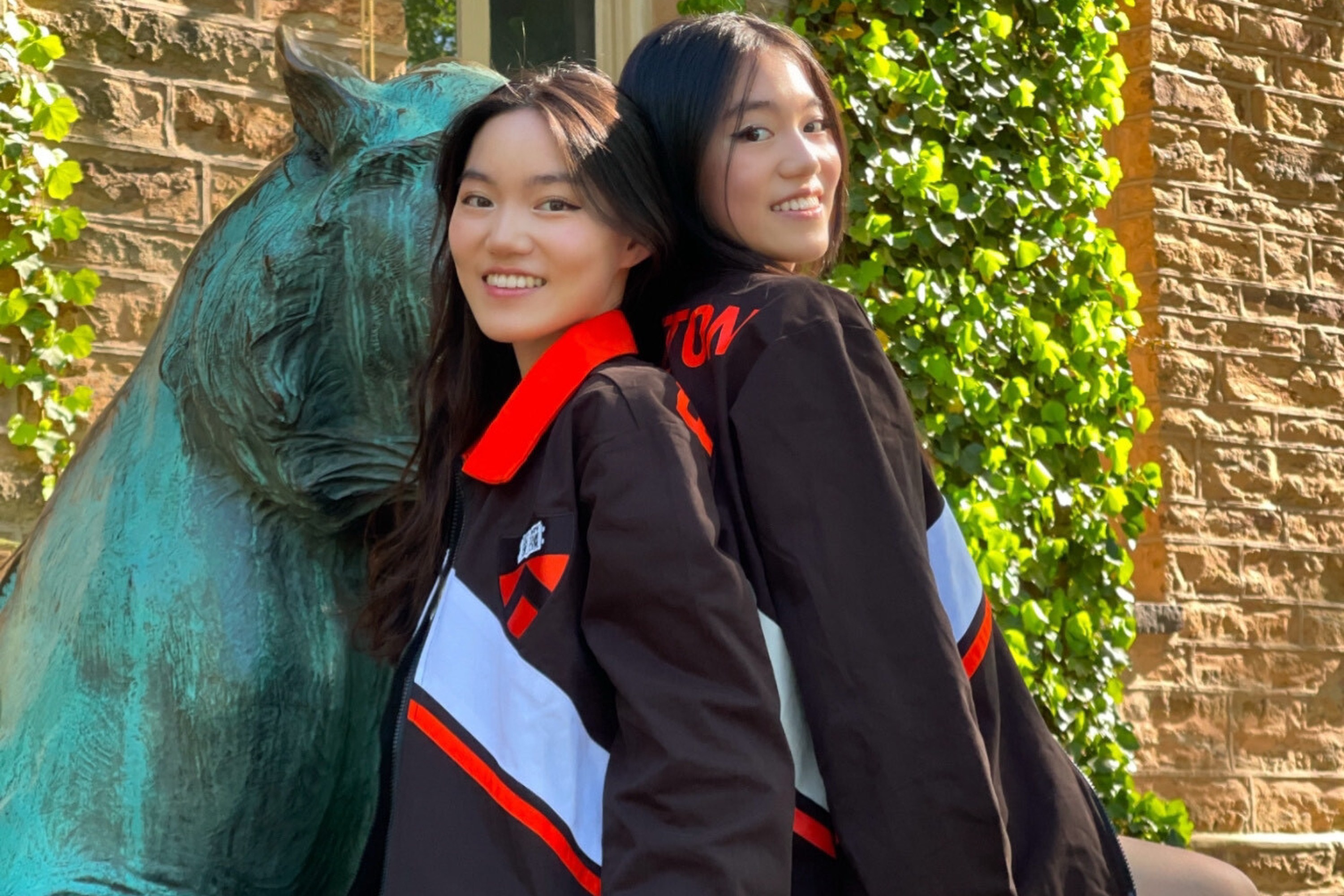 Two identical twins smile and stand back to back in matching jerseys in front of a lion statue.