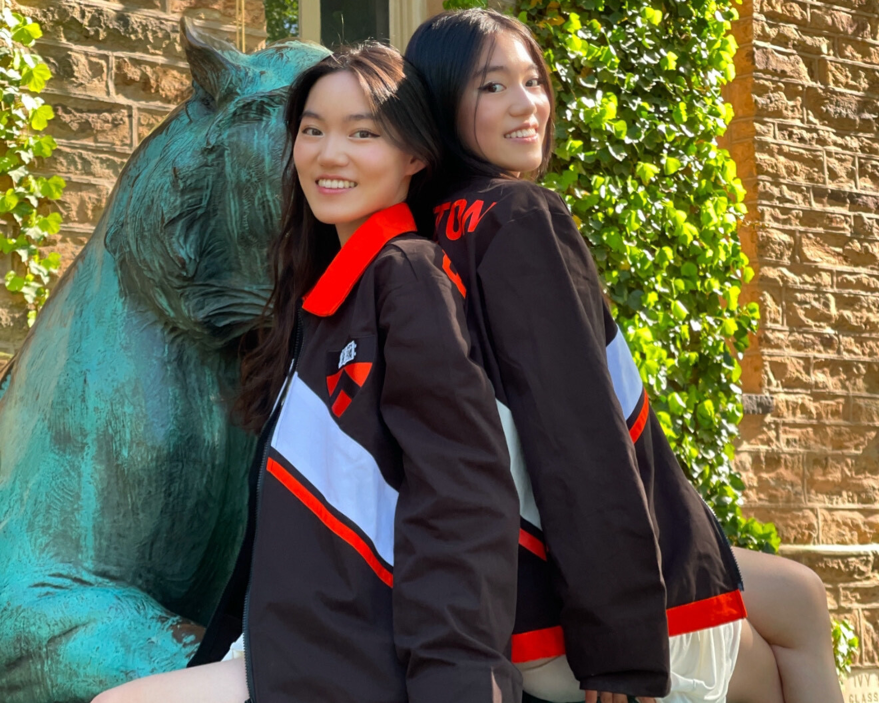 Two identical twins smile and stand back to back in matching jerseys in front of a lion statue.