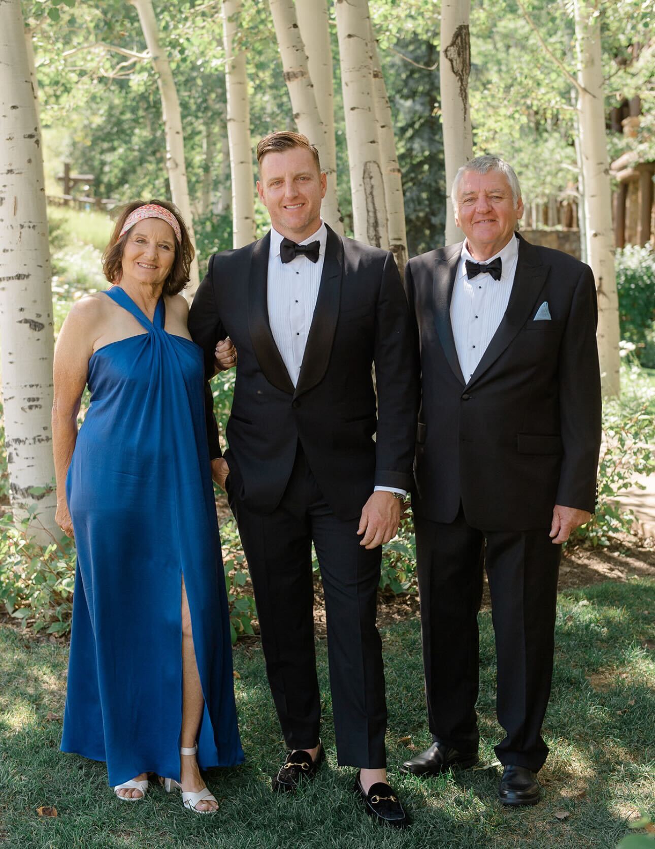 Three people are posing in a lush, sunlit grove with white-barked trees. The woman wears a blue dress, and both men are in black tuxedos.
