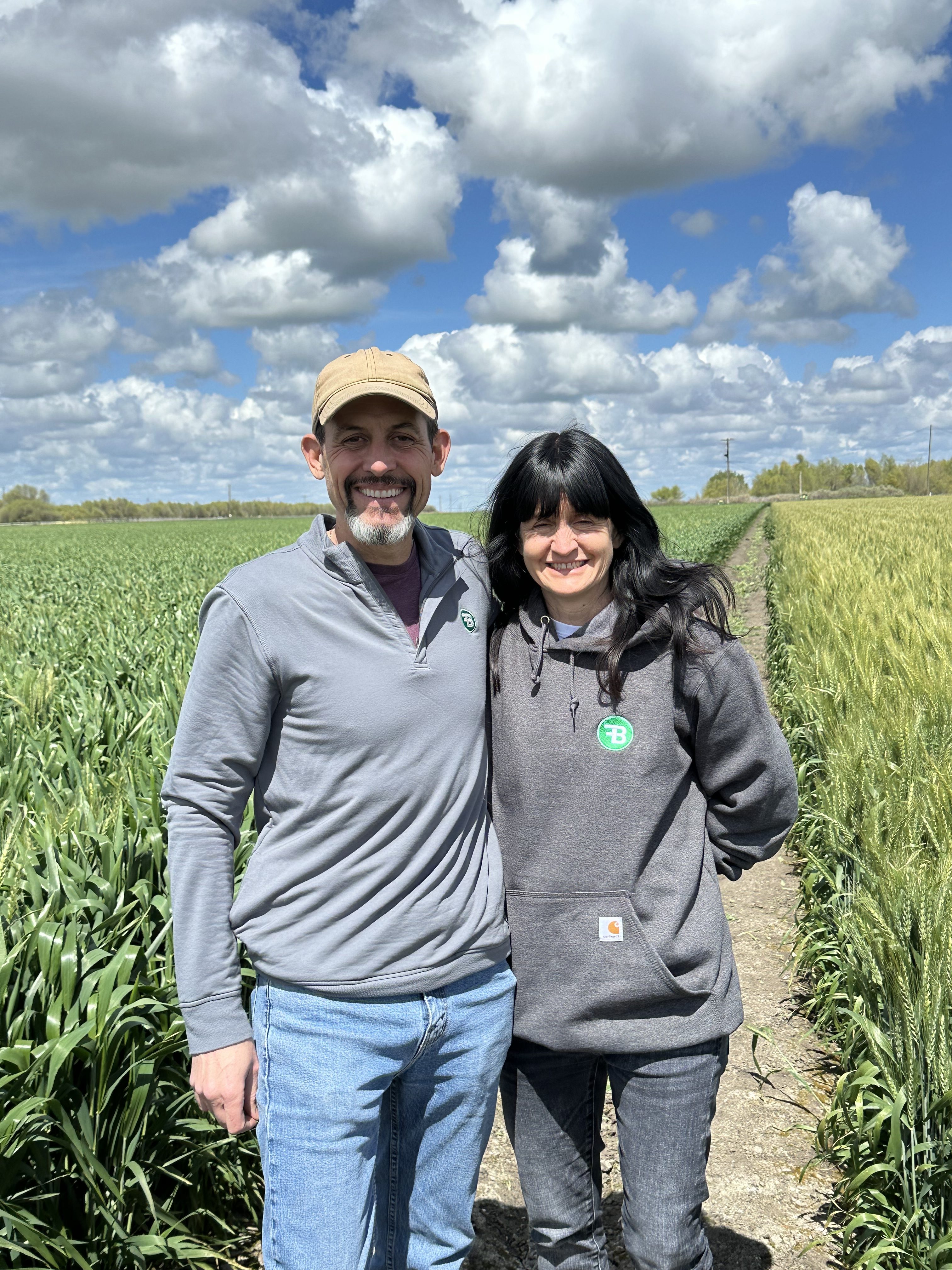 A smiling couple stands side by side on a path through lush green fields, beneath a sky filled with fluffy clouds. Both wear casual clothes and look content.