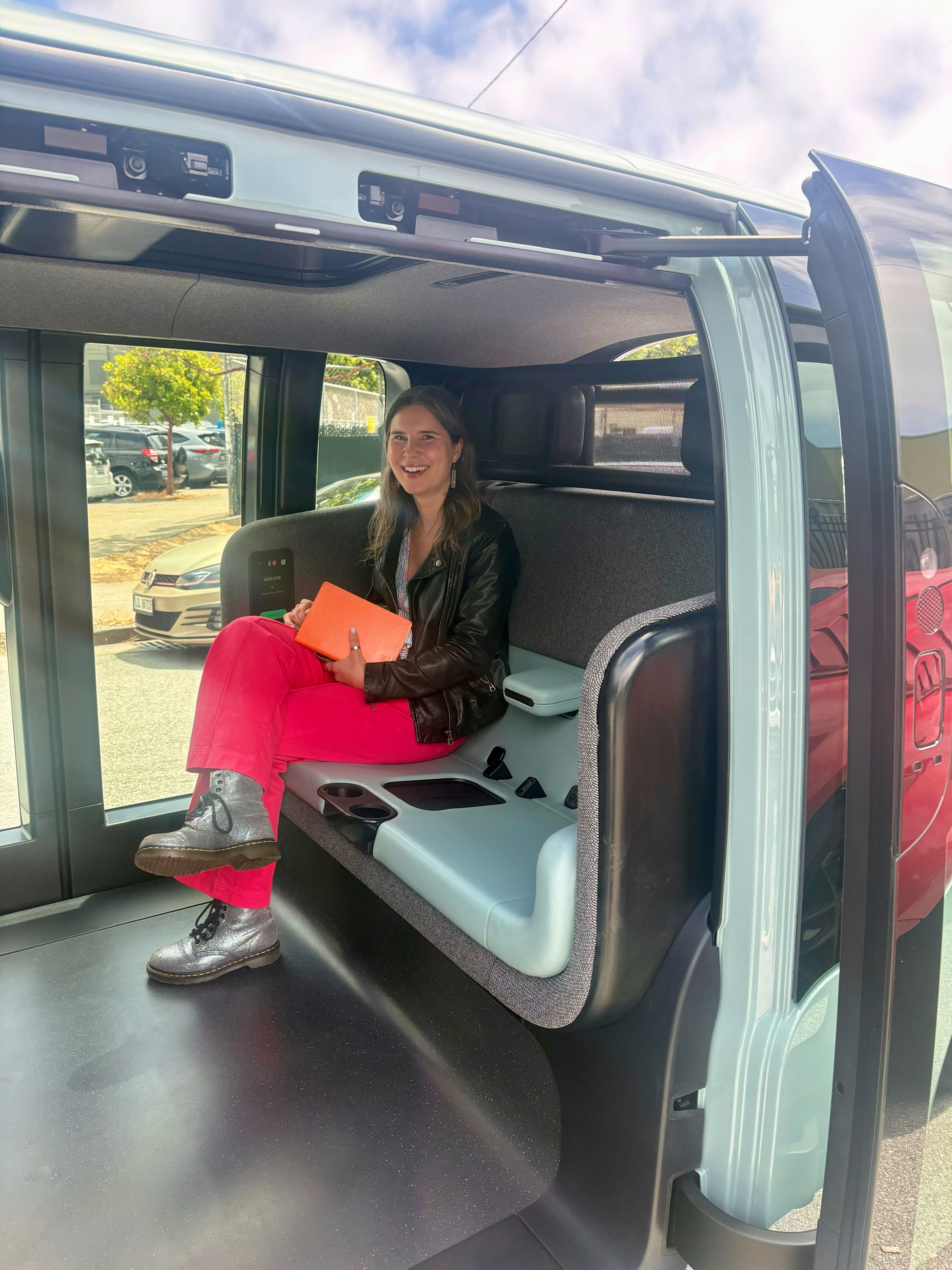 A person sits smiling inside a sleek vehicle with open sliding doors. They wear a leather jacket, red pants, shiny boots, and hold an orange tablet.