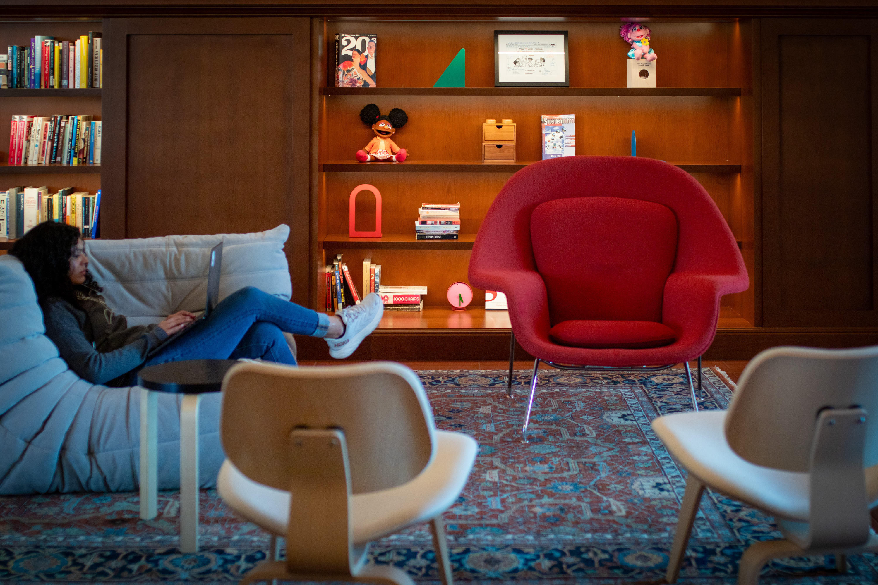 A cozy room with bookshelves, a person on a light blue sofa with a laptop, a red armchair, wooden chairs, and a patterned rug. Shelves have books and decorations.