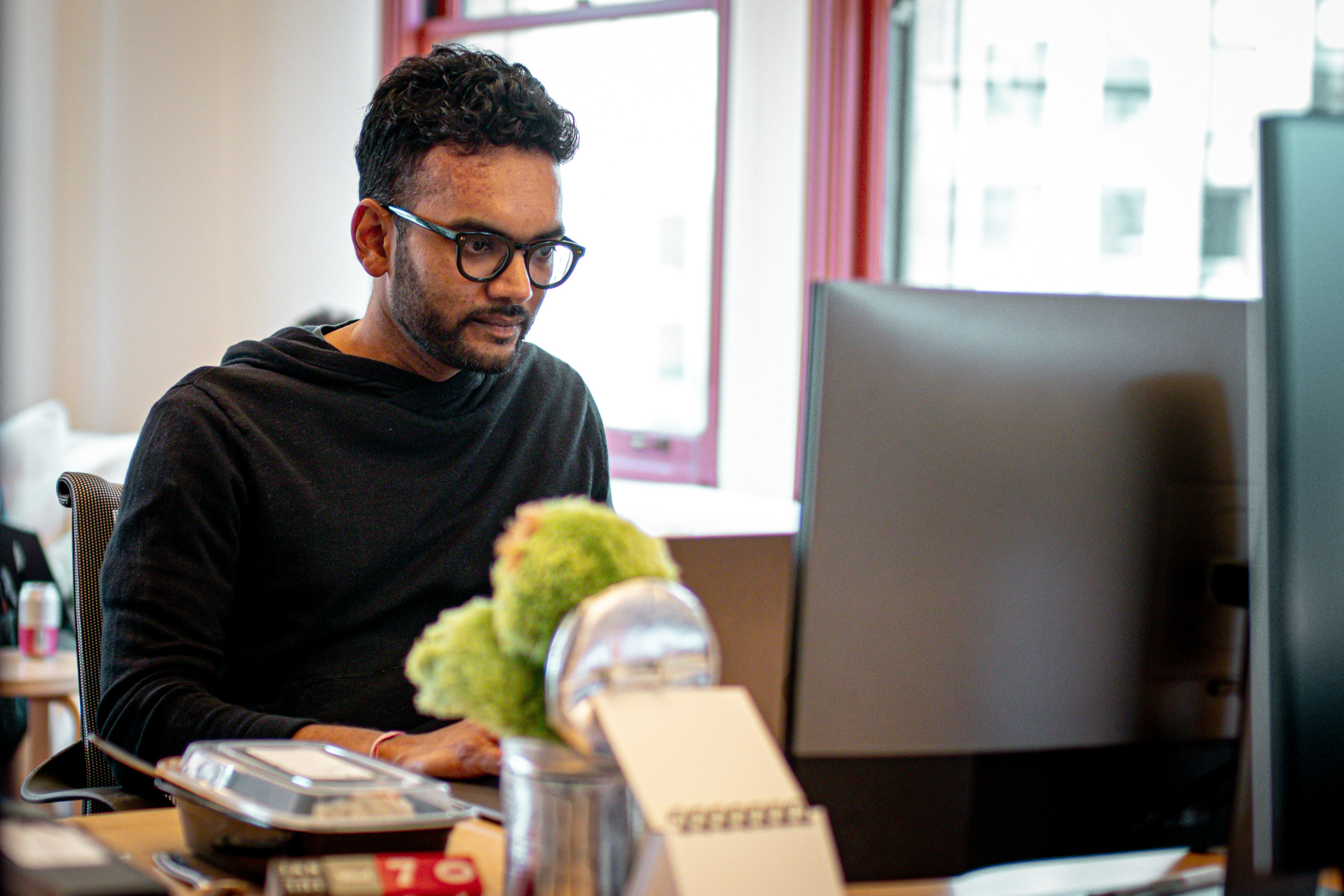 A man with glasses and dark hair is focused on his computer at a desk. There's a notebook, takeaway container, and a tin holding green objects on the desk.