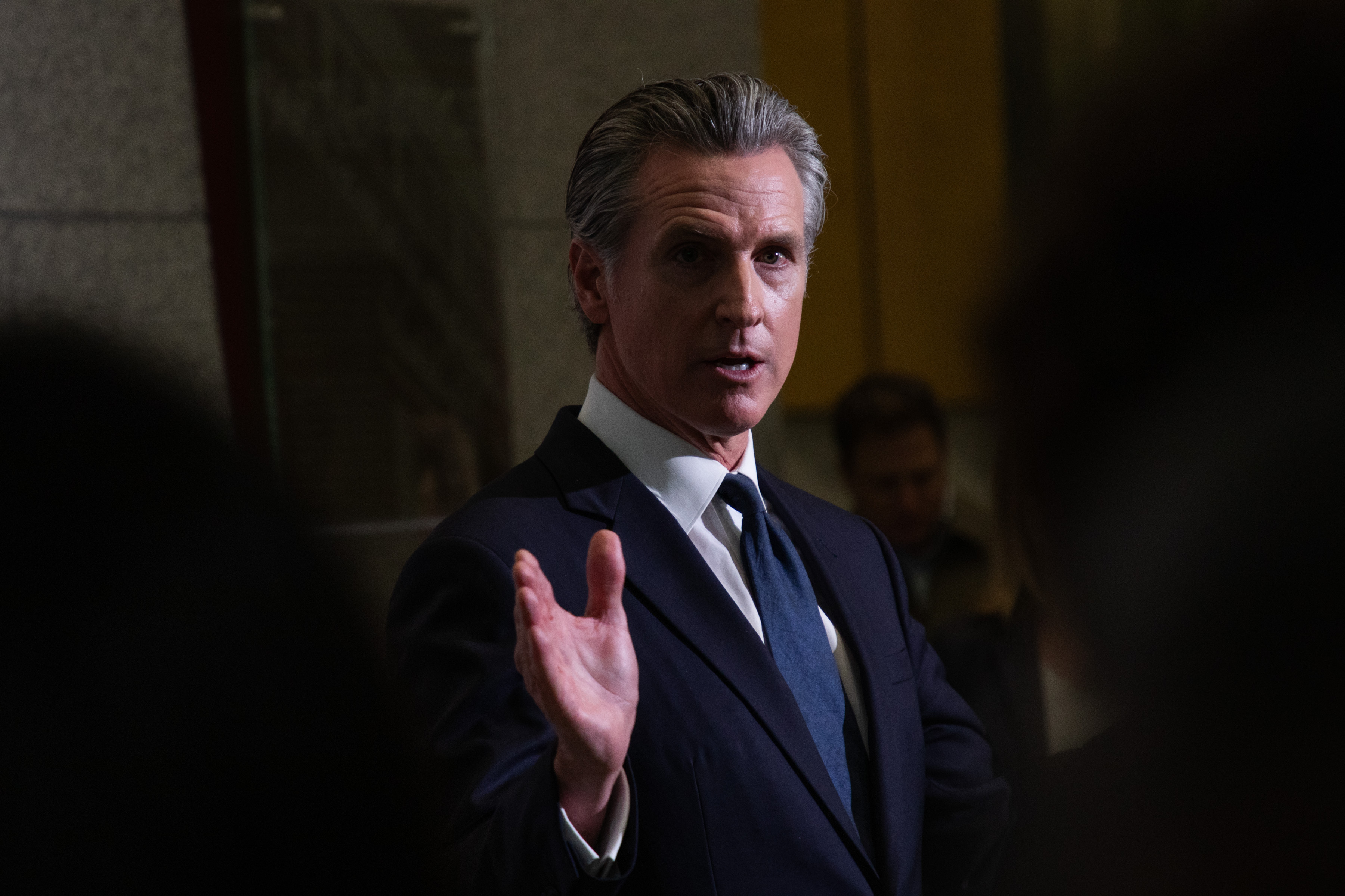 A man in a dark suit gestures with one hand raised slightly. He is standing indoors, lit by soft, focused lighting with a few blurred people in the background.
