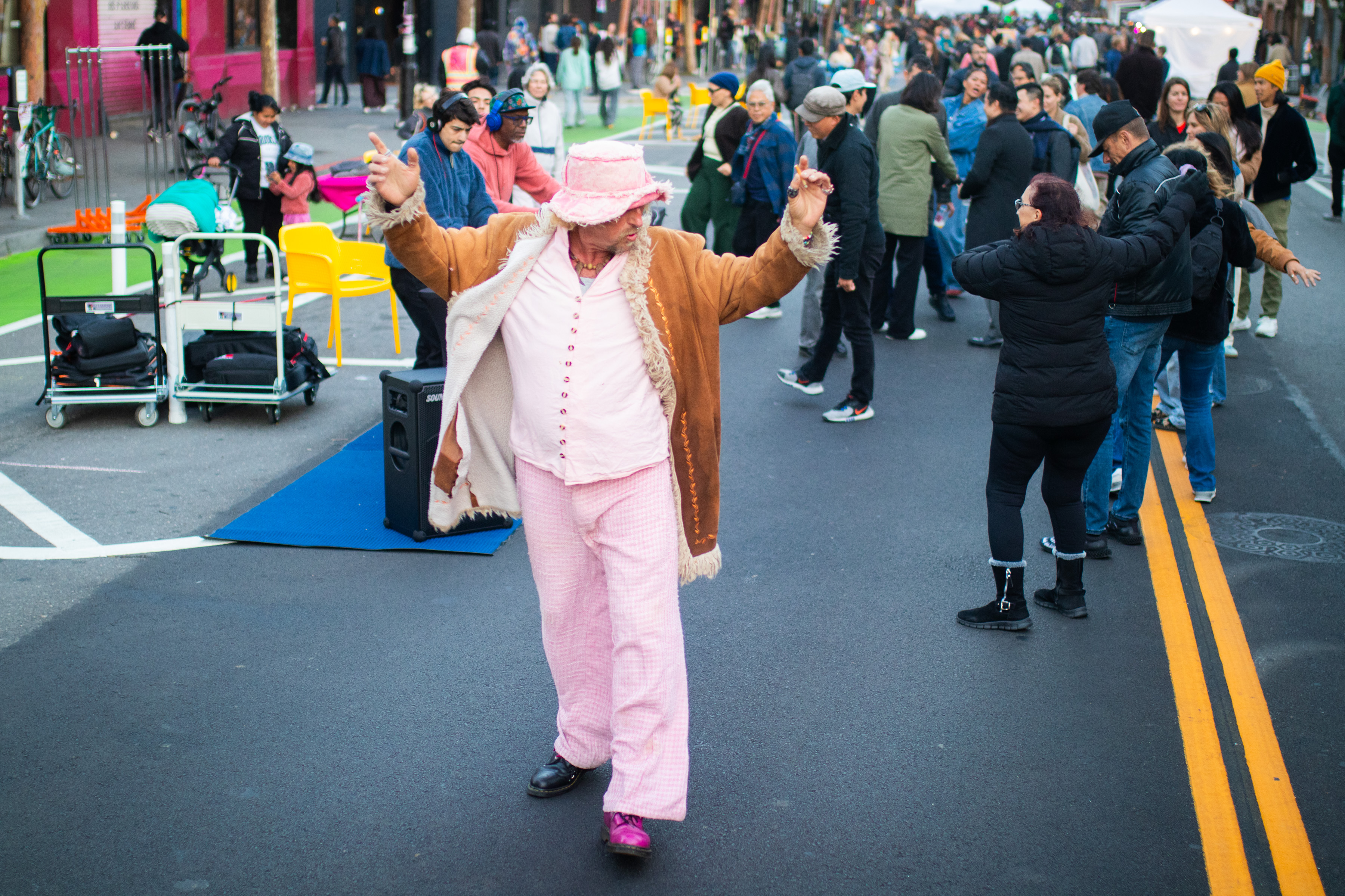 A person in a pink outfit and hat dances joyfully on a street surrounded by a crowd of people. The atmosphere is lively, with onlookers and street activity.