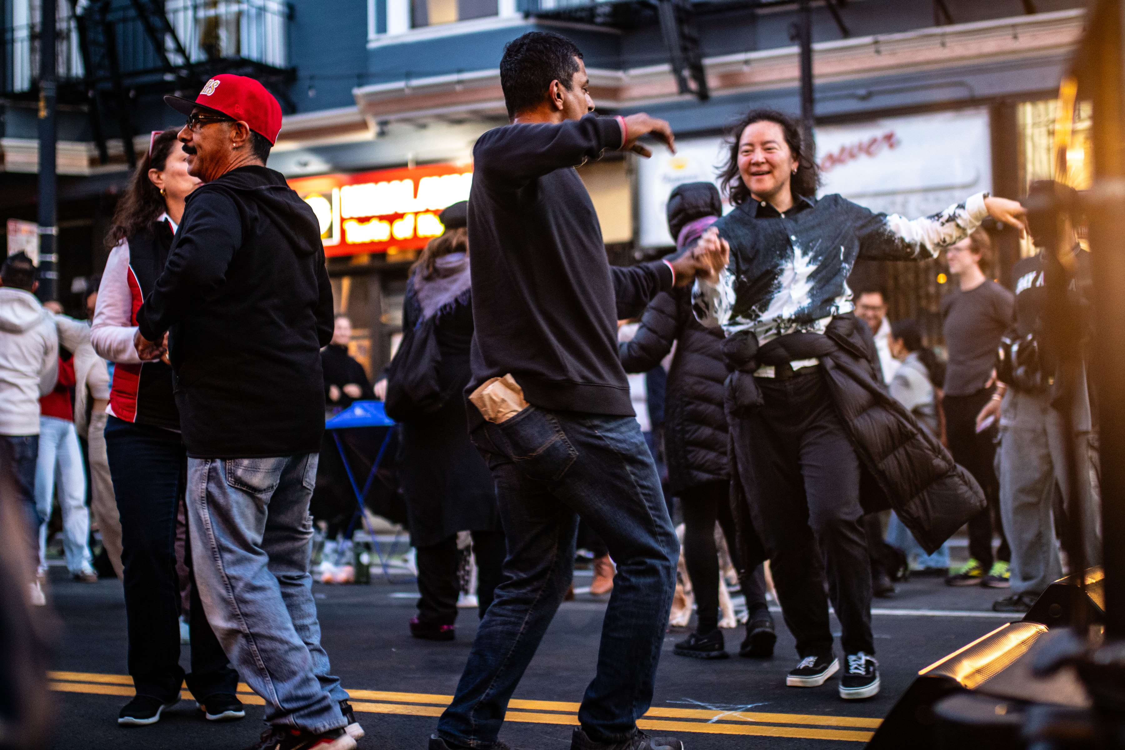 A lively street scene features people joyfully dancing. The crowd appears relaxed, with some individuals smiling and holding hands. The background is an urban setting.