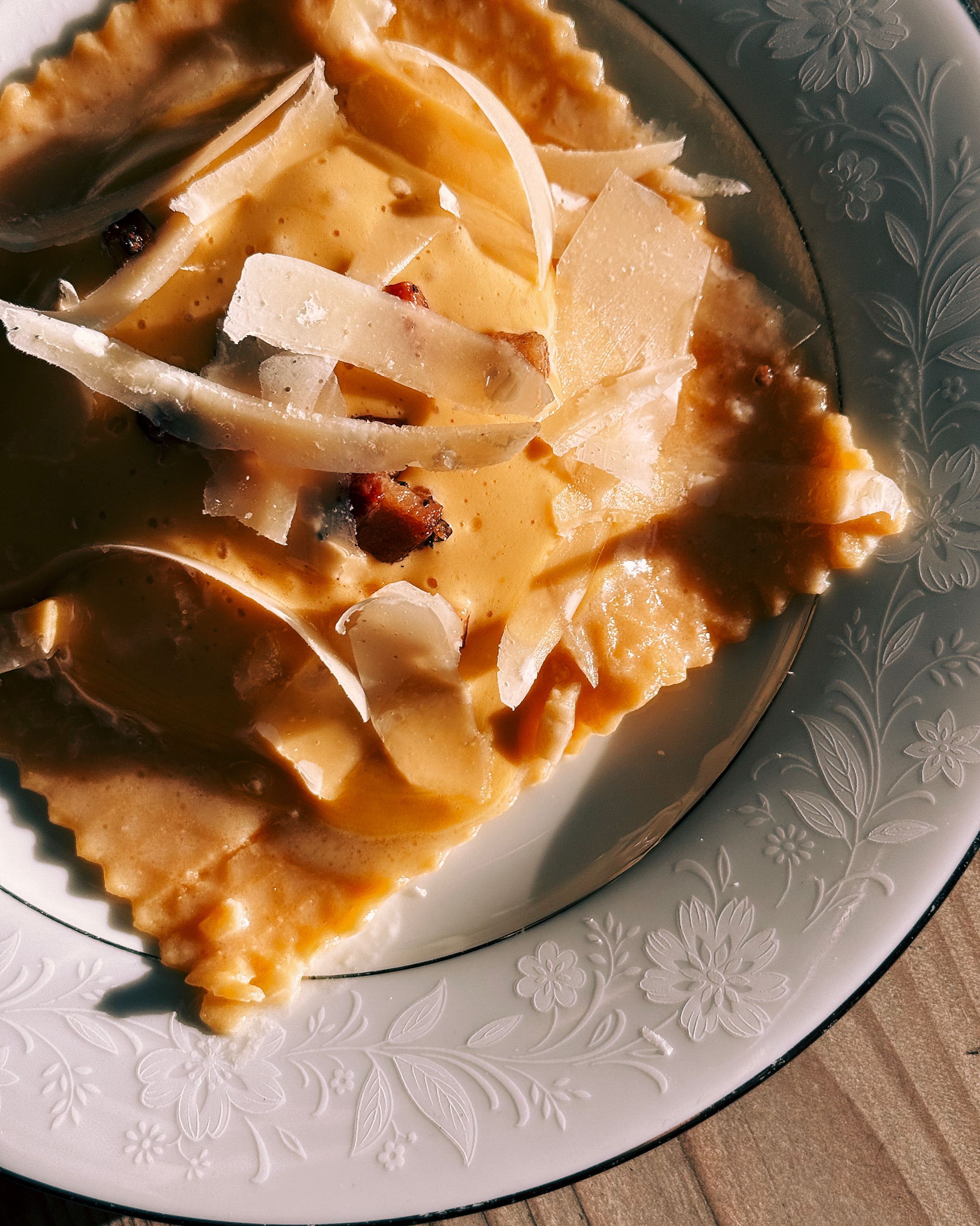 A plate with ravioli topped with creamy sauce, shaved cheese, and small bacon pieces, rests on a floral-patterned dish.