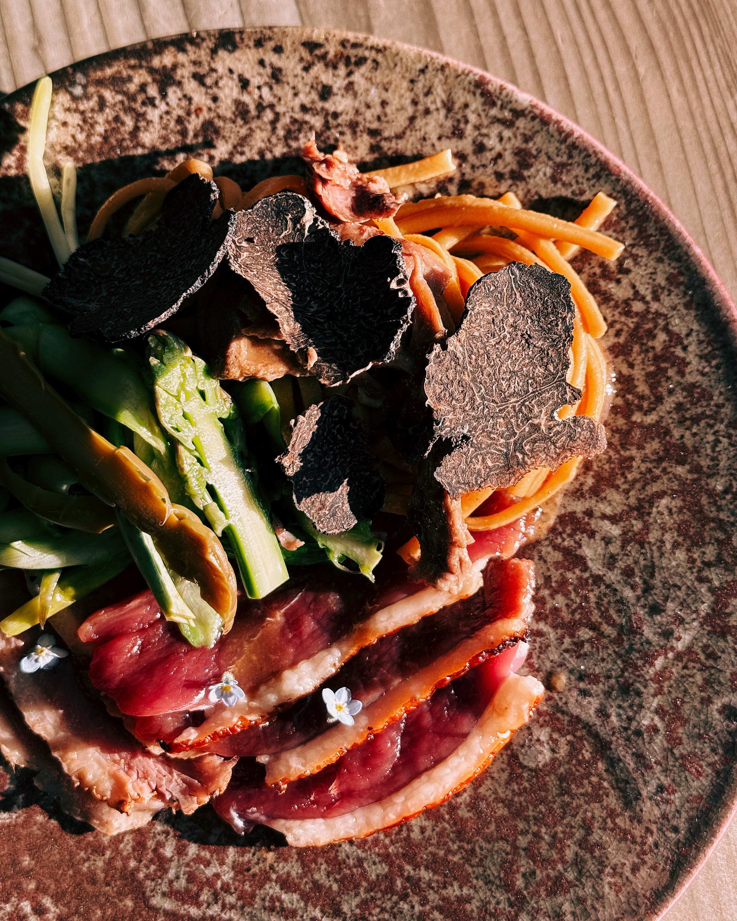 The image shows a plated dish with thinly sliced meat, asparagus, pasta, and garnished with black truffle slices and small white flowers.