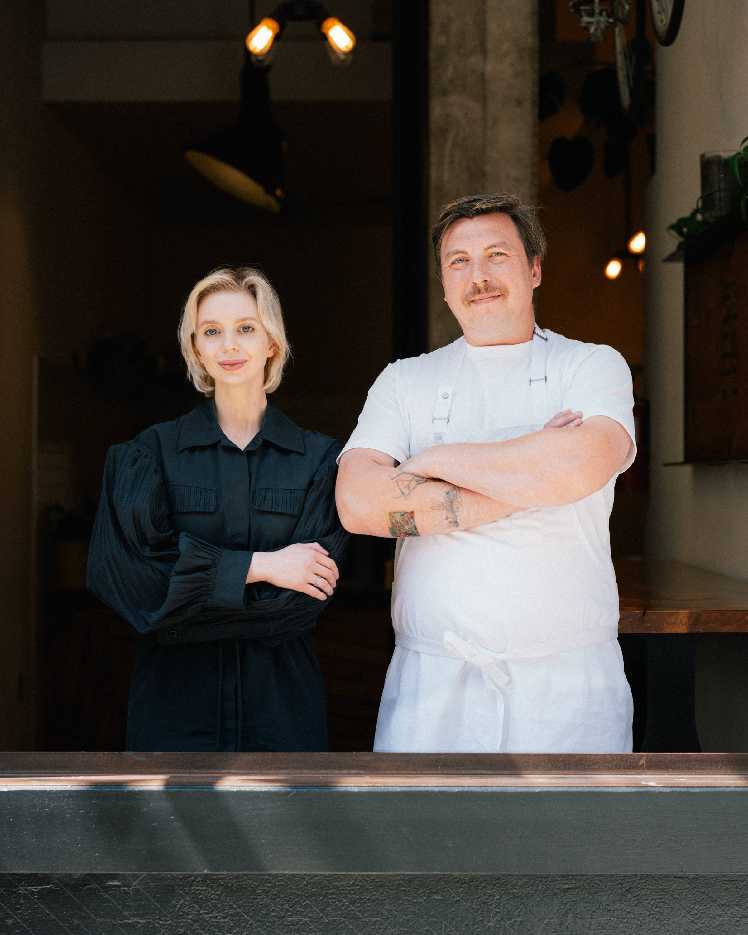 A man in a white shirt and apron stands beside a woman in a black dress. They're in a dimly lit space with light fixtures above them, appearing calm and composed.