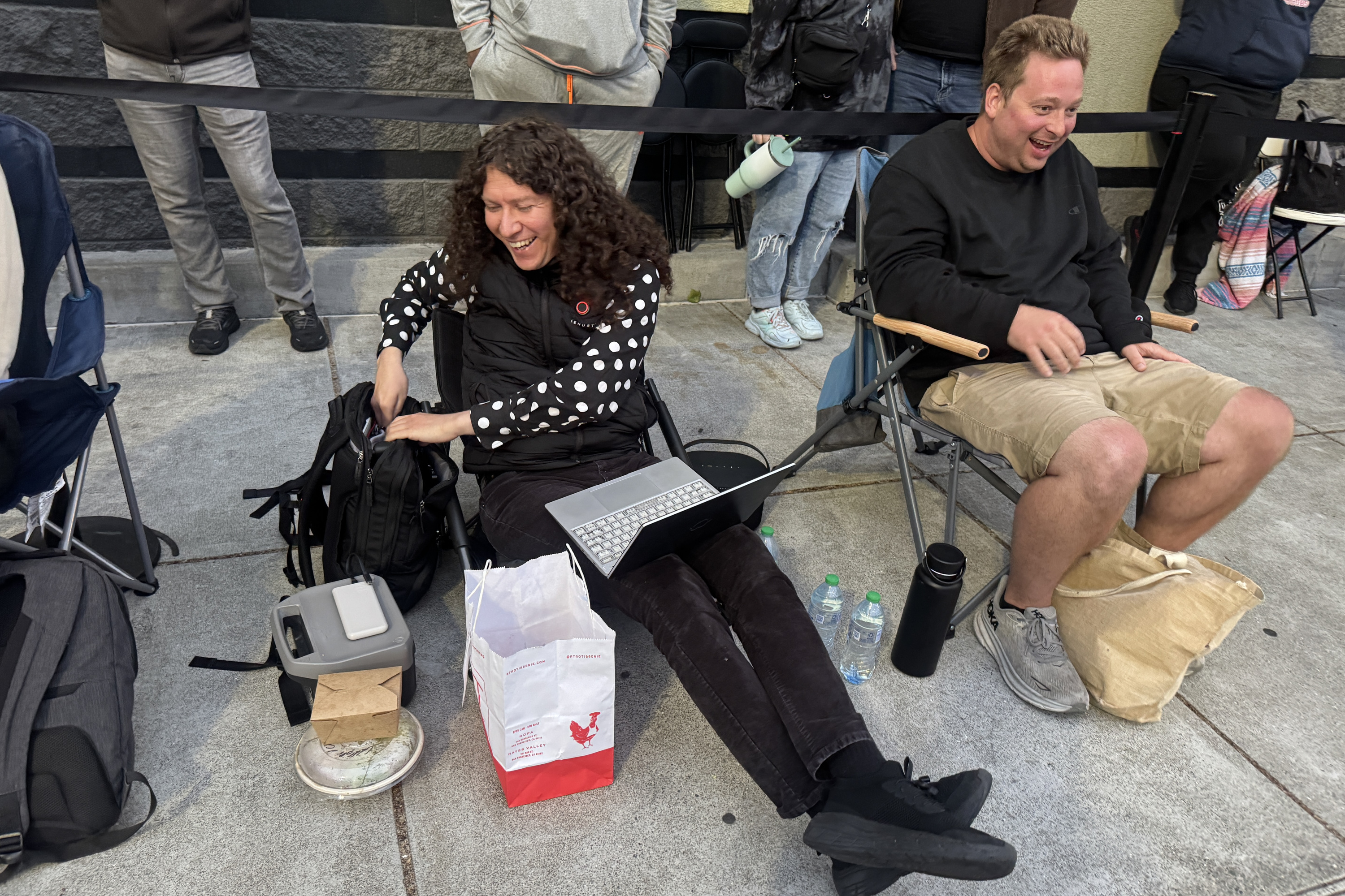 Two people are sitting on folding chairs on a sidewalk. The person on the left is using a laptop, while both are smiling. Bottled water and food bags are beside them.