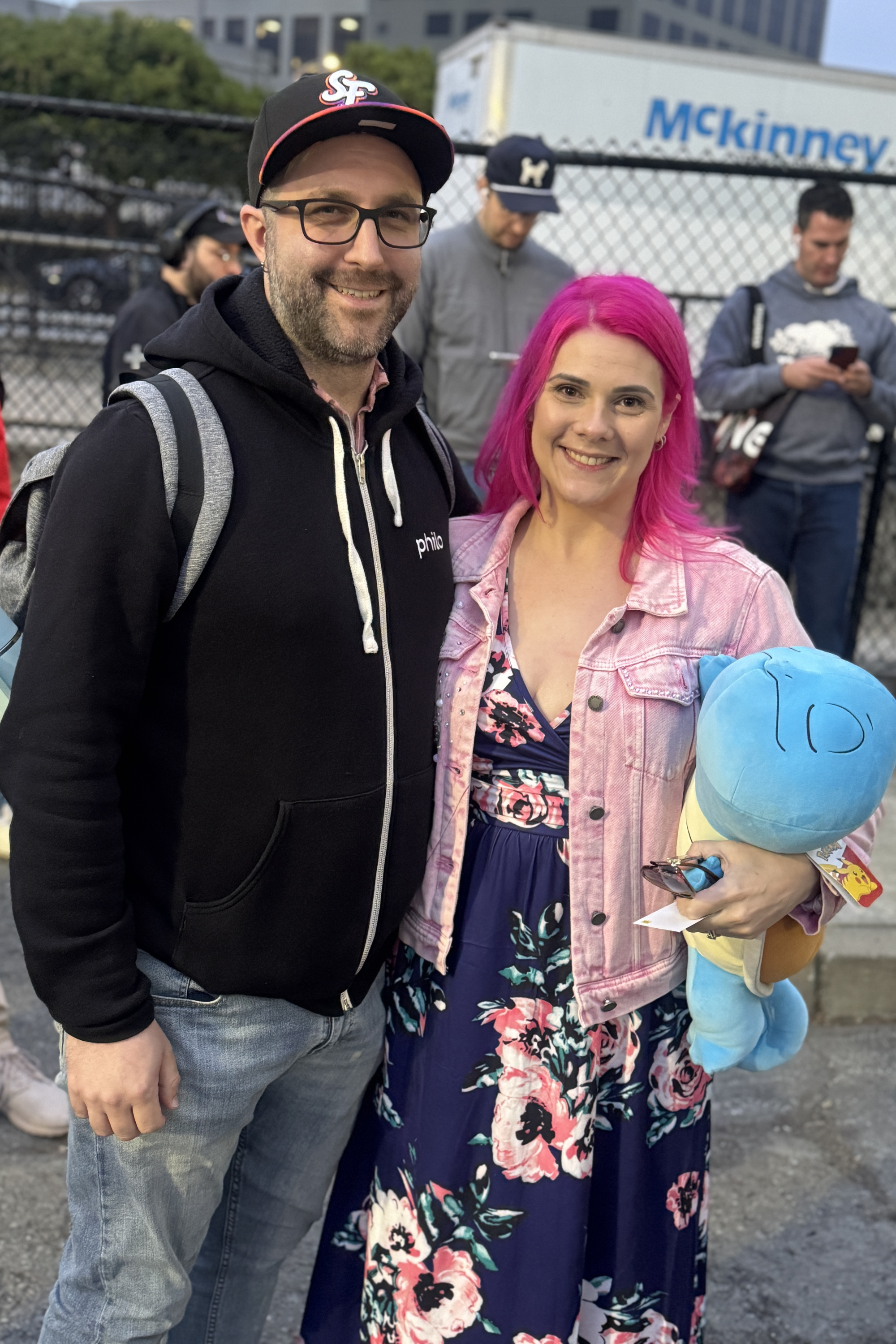 A man in a black hoodie and cap stands with a woman in a pink jacket holding a blue plush toy. She has vibrant pink hair and wears a floral dress.
