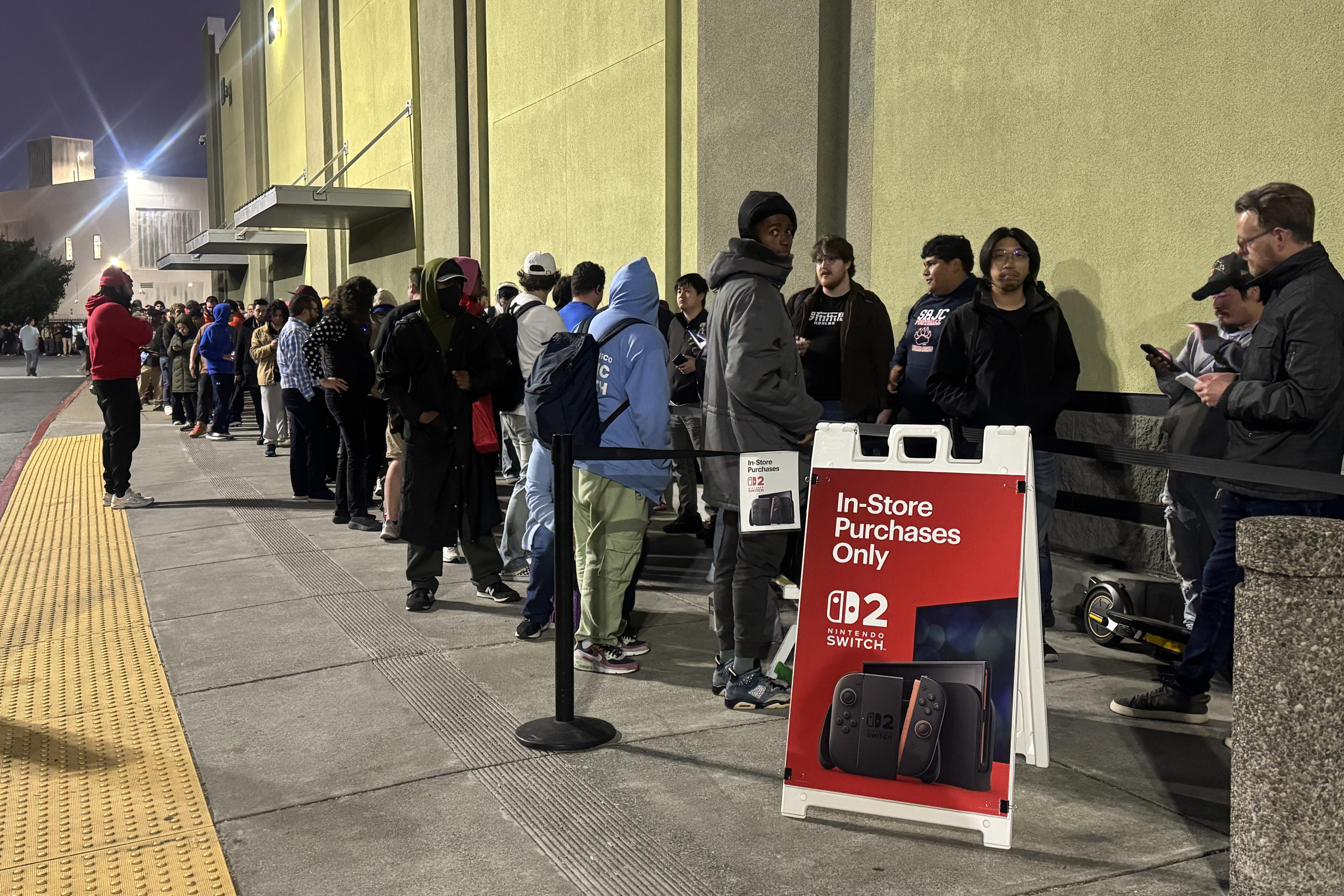 A crowd of people in jackets line up outside a store at night. A red sign reads &quot;In-Store Purchases Only&quot; for the &quot;Nintendo Switch 02.&quot;