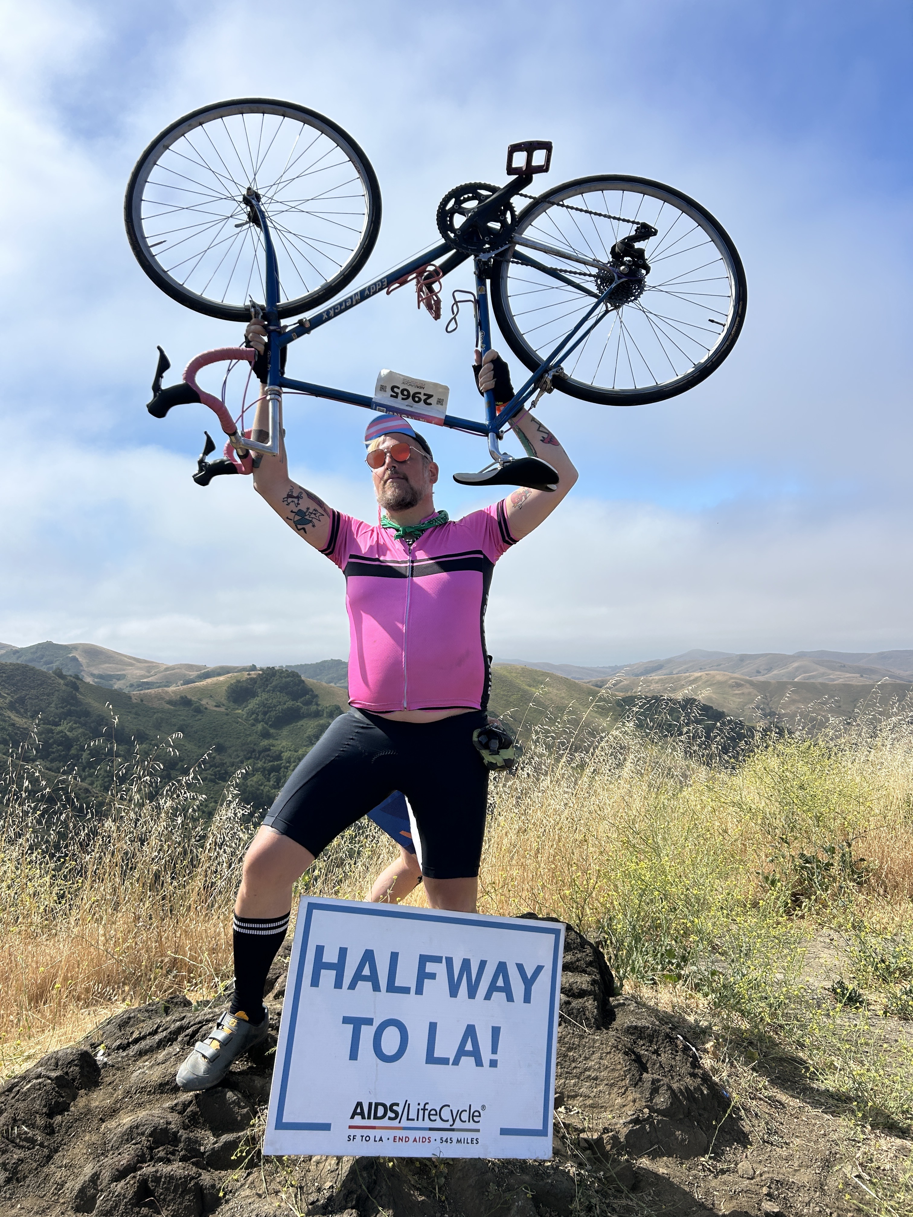 A cyclist in a pink jersey holds a bike overhead triumphantly on a hill. A sign reads &quot;HALFWAY TO LA! AIDS/LifeCycle&quot; against a backdrop of rolling hills.