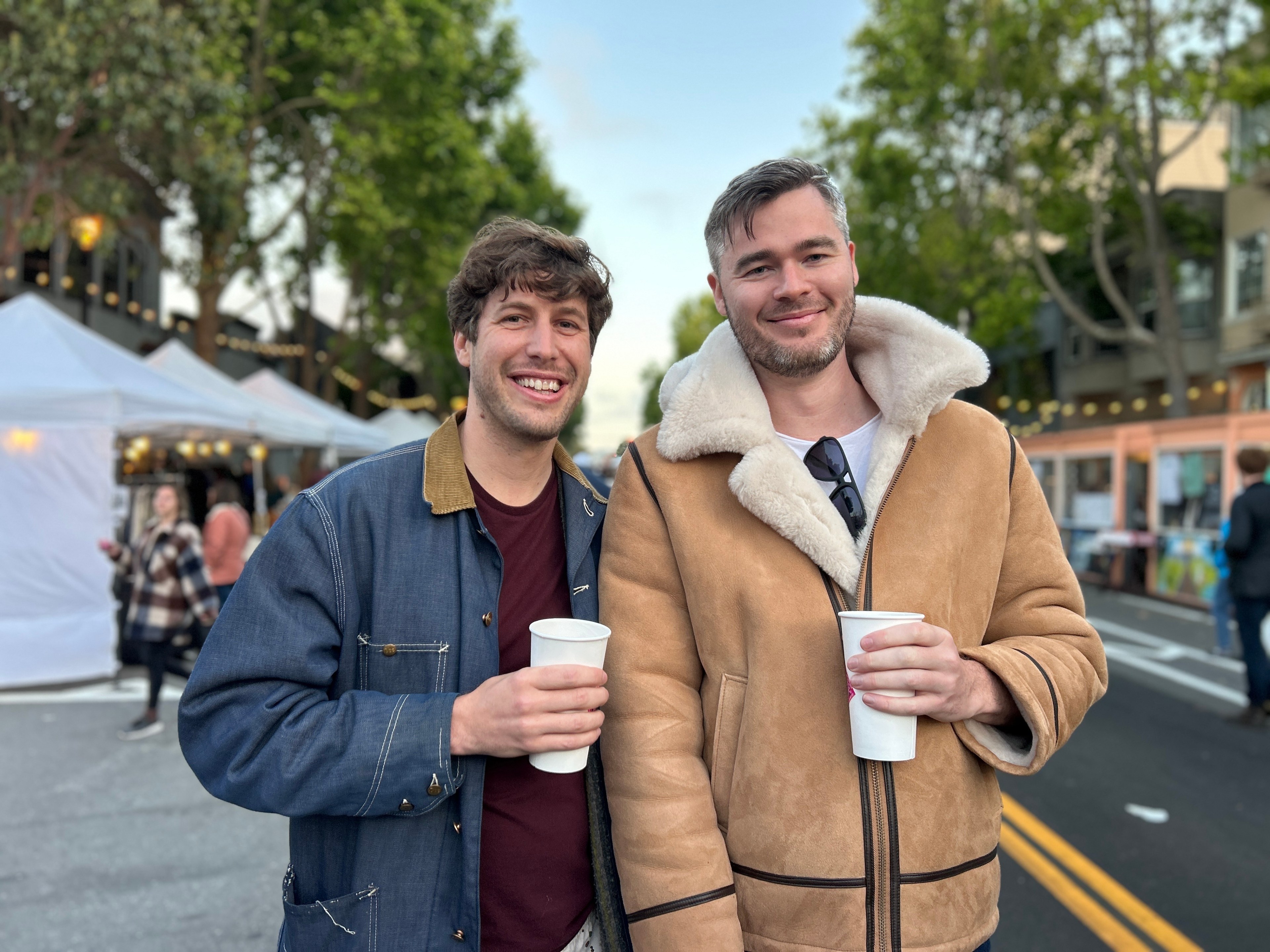 Two people stand smiling on a street lined with tents and trees. They are holding cups and wearing jackets, one blue and the other tan with a fuzzy lining.