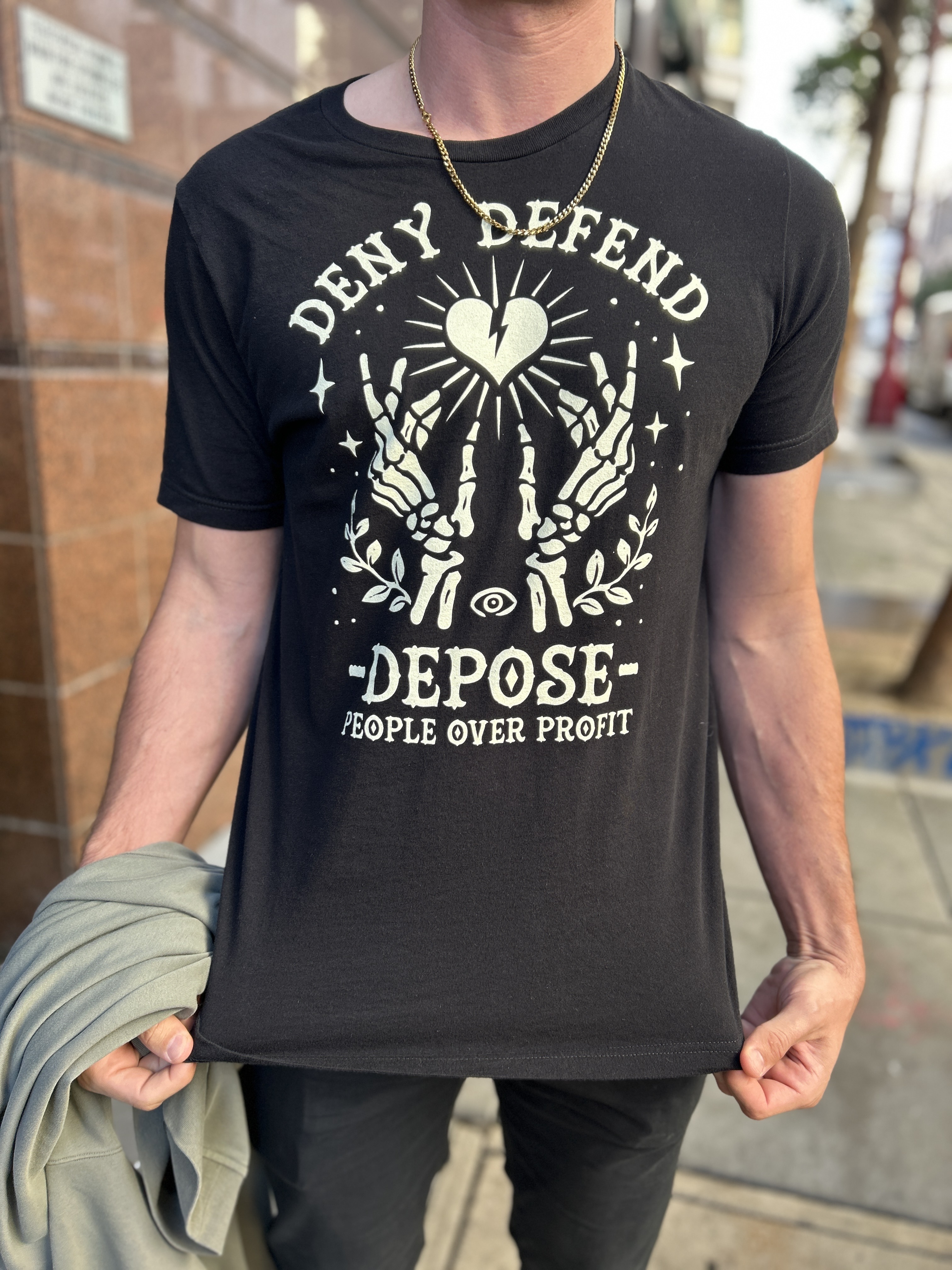 A person wears a black T-shirt with the words "Deny Defend Depose" and "People Over Profit" in bold. The design includes skeletal hands, a broken heart, and an eye.
