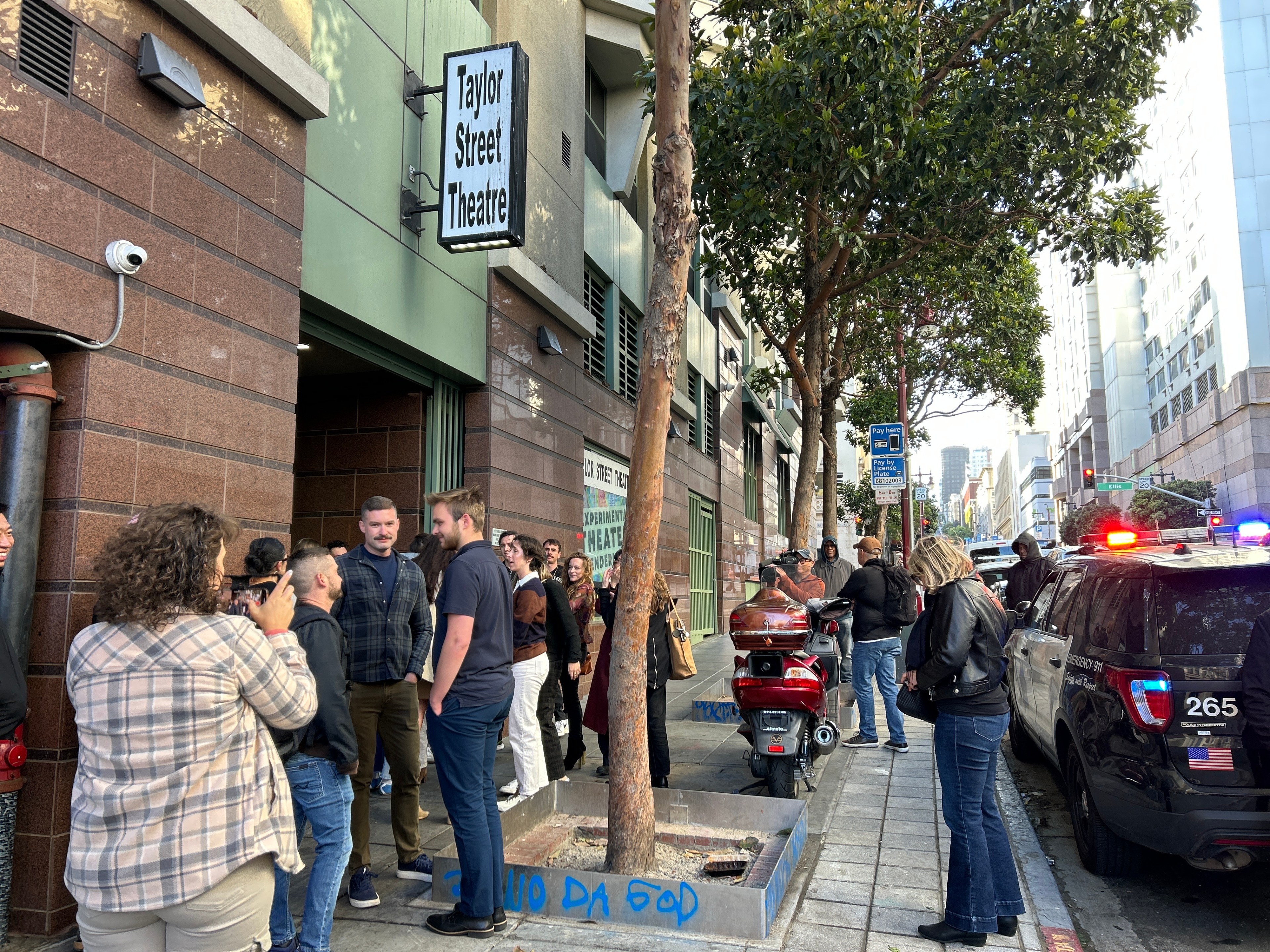 A crowd gathers outside Taylor Street Theatre. People chat near the entrance while a police car with flashing lights is parked on the street.