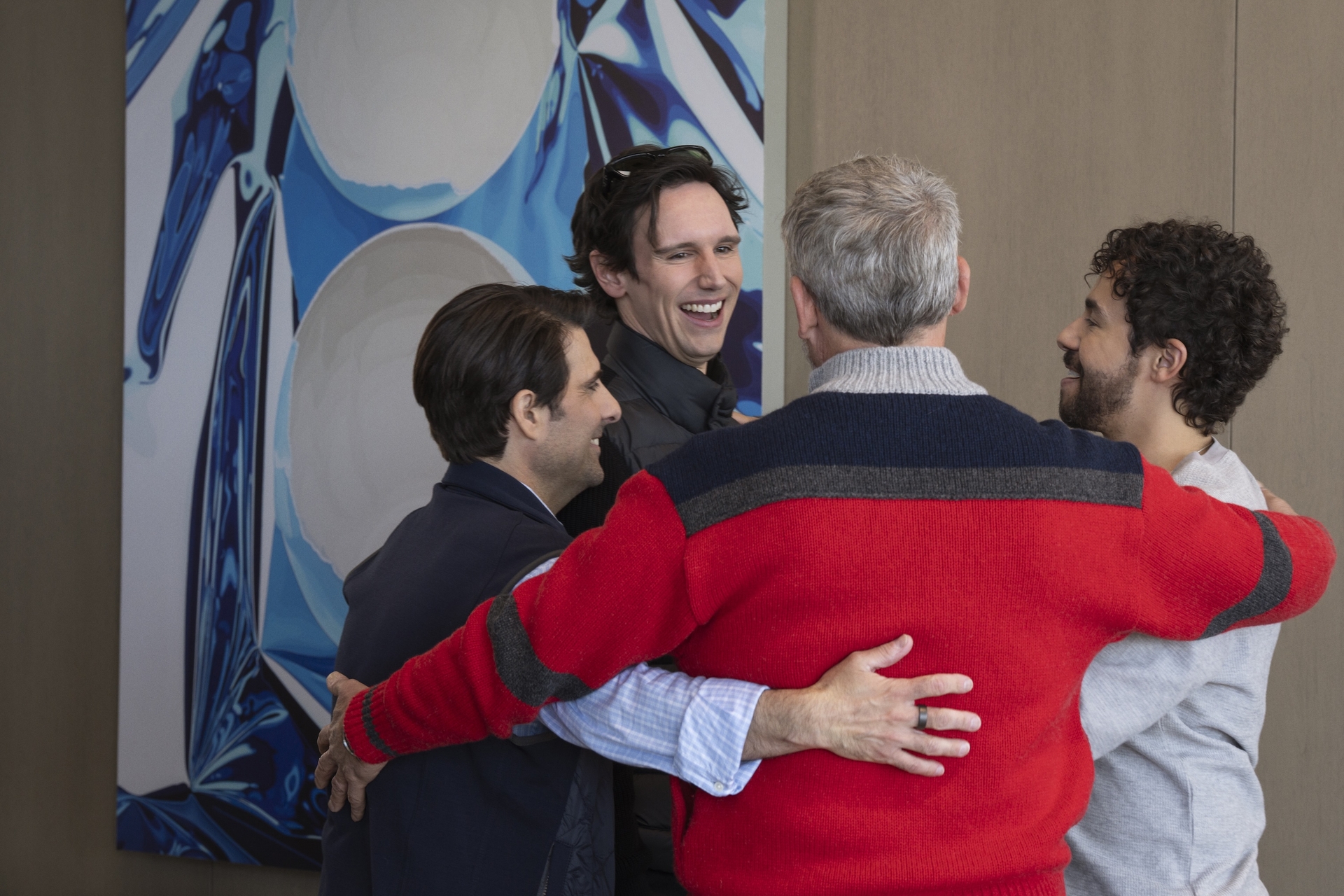 Four men stand in a group hug, smiling and laughing. They are in front of a large abstract painting with blue and white colors.