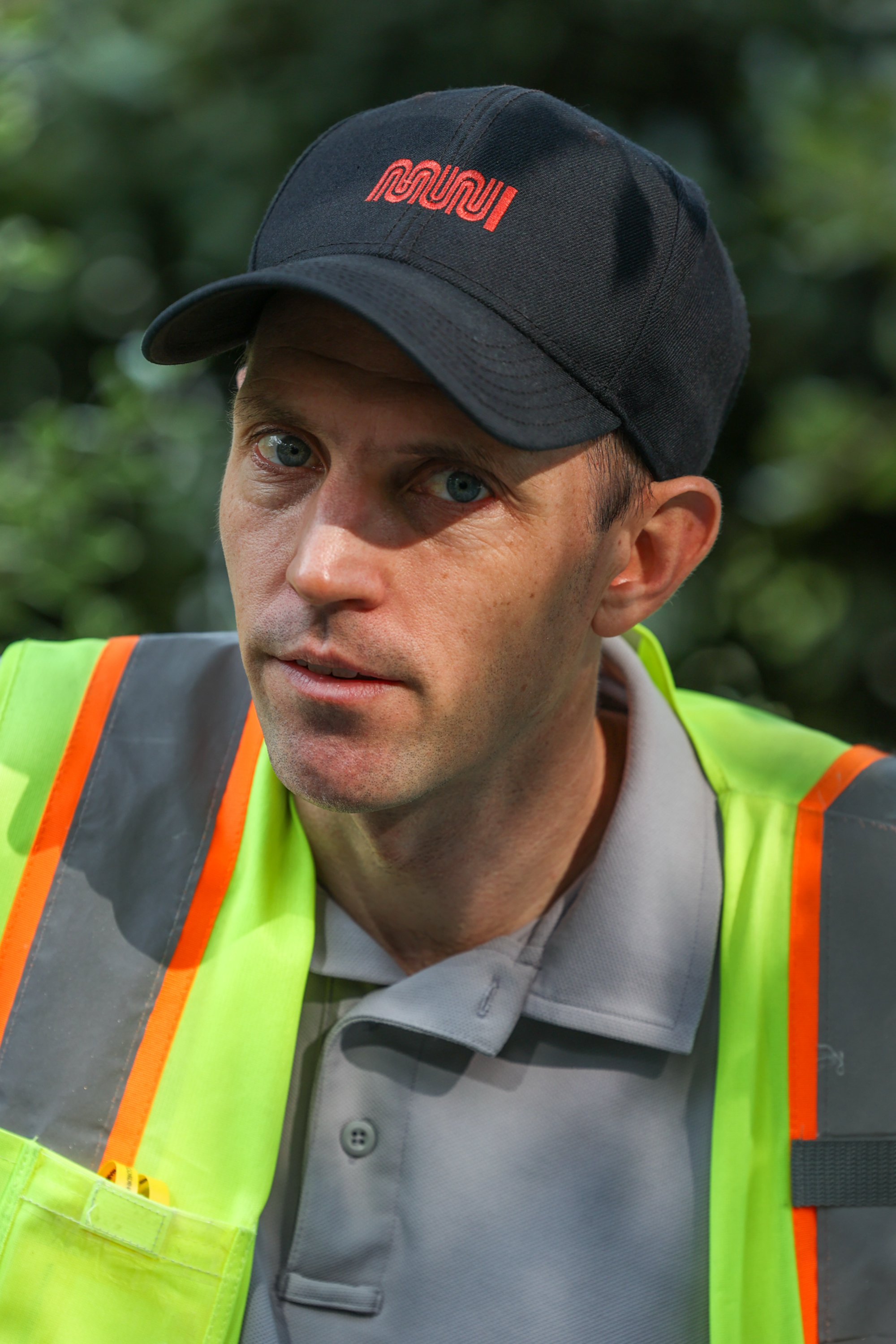 A person wearing a black cap with red text and a neon yellow safety vest with orange stripes looks directly at the camera in a natural outdoor setting.
