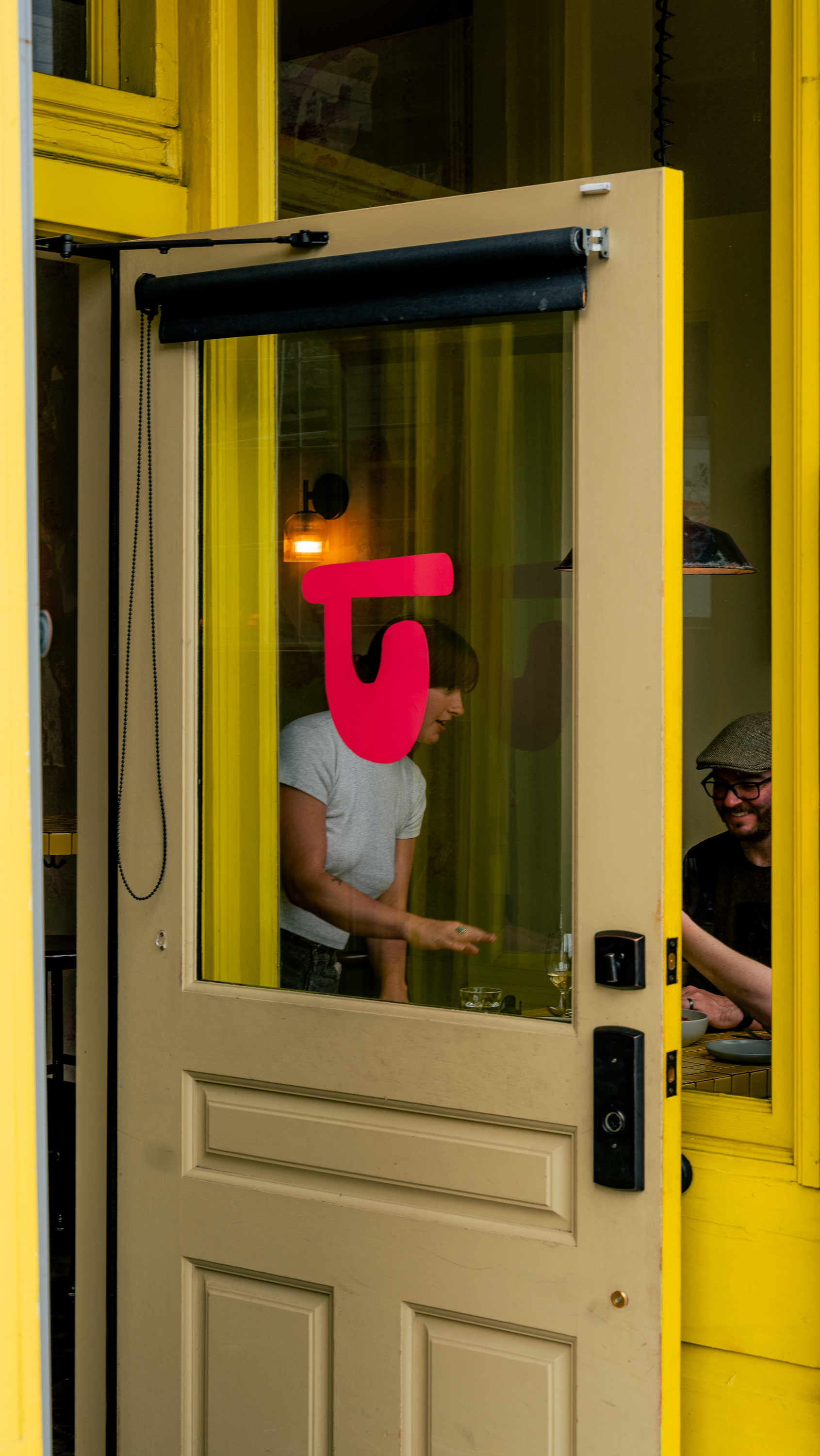 A partially open yellow door with a pink logo shows two people inside, one gesturing, another smiling, amidst a warm-lit interior.