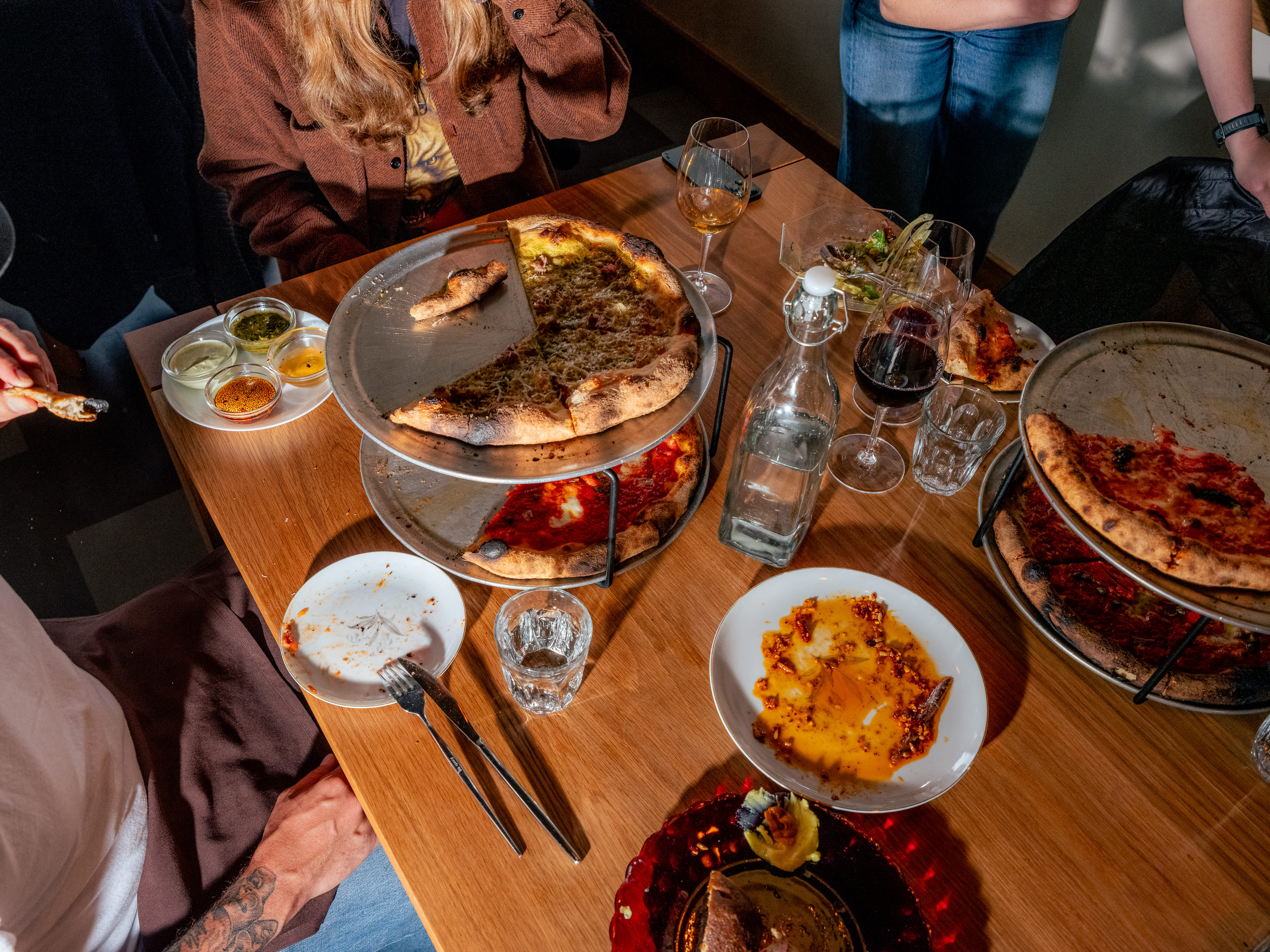 A table with half-eaten pizzas on metal stands, glasses of wine, an empty plate, and sauce dishes. People are seated and standing around, enjoying the meal.