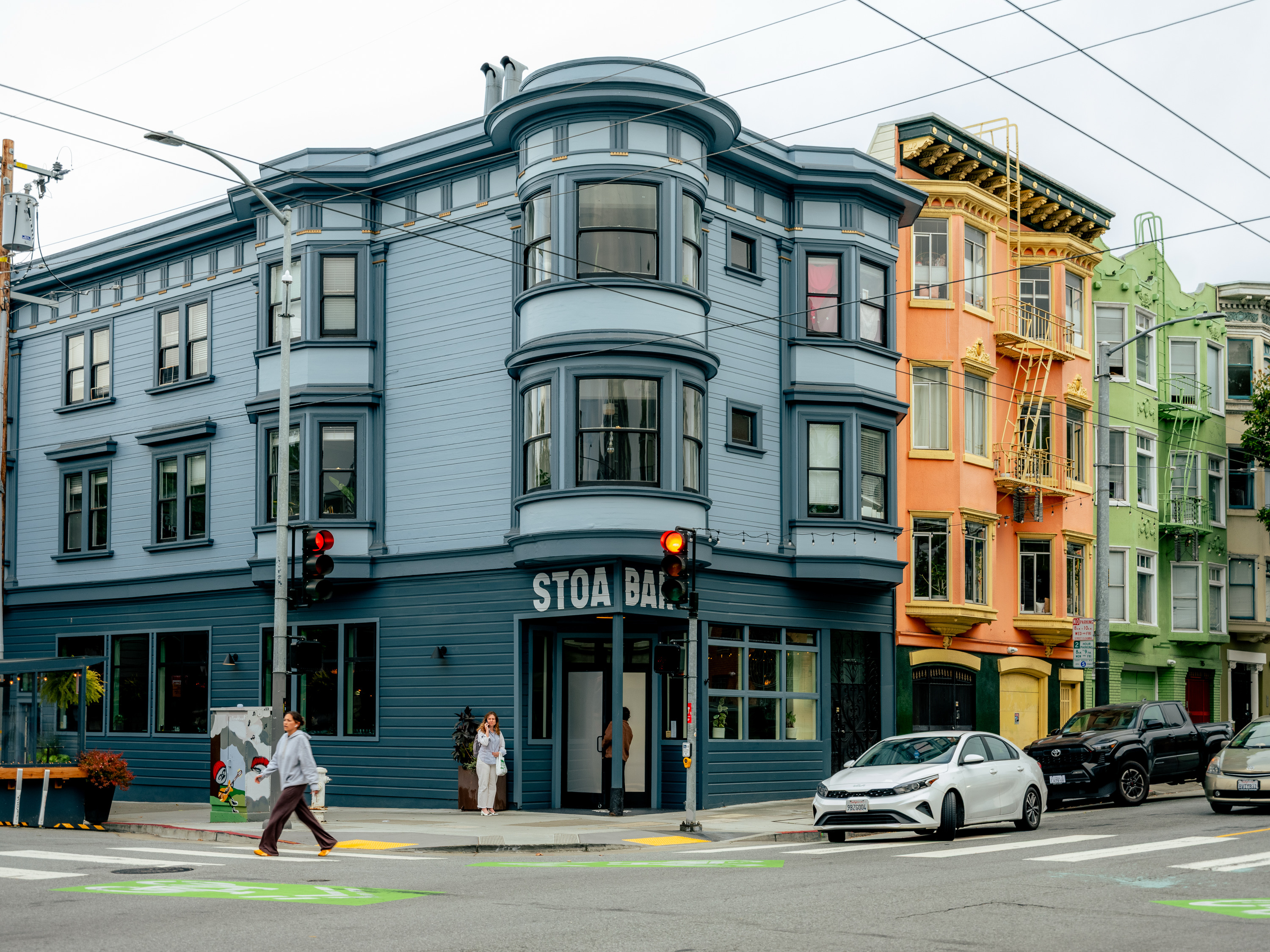 A street corner features colorful buildings: a blue one with arched windows, an orange building, and a green one. A person crosses the street, while cars wait.