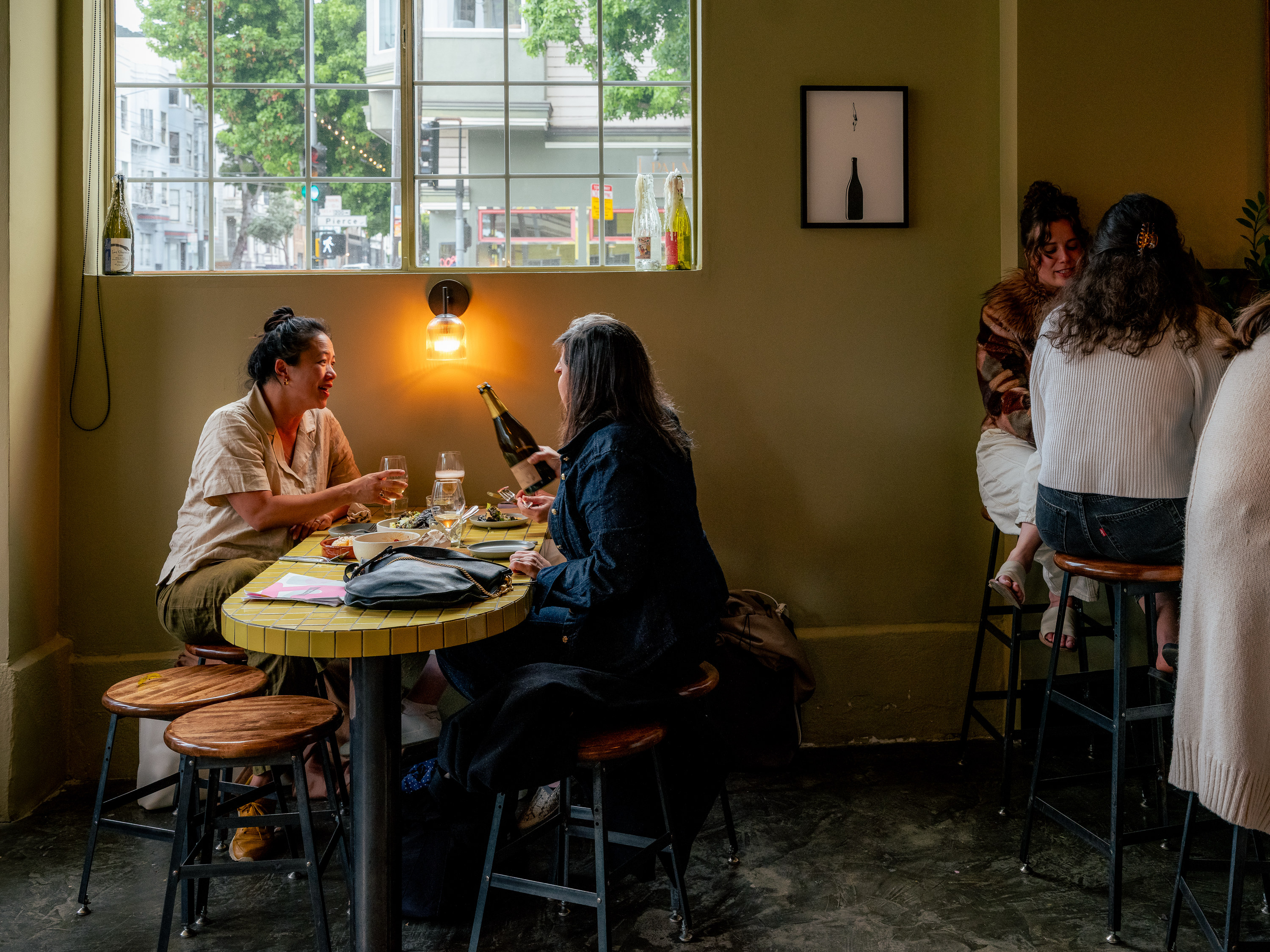 Two people sit at a cozy cafe table with a bottle and glasses, engaged in conversation. Warm light creates an inviting atmosphere, with others nearby.