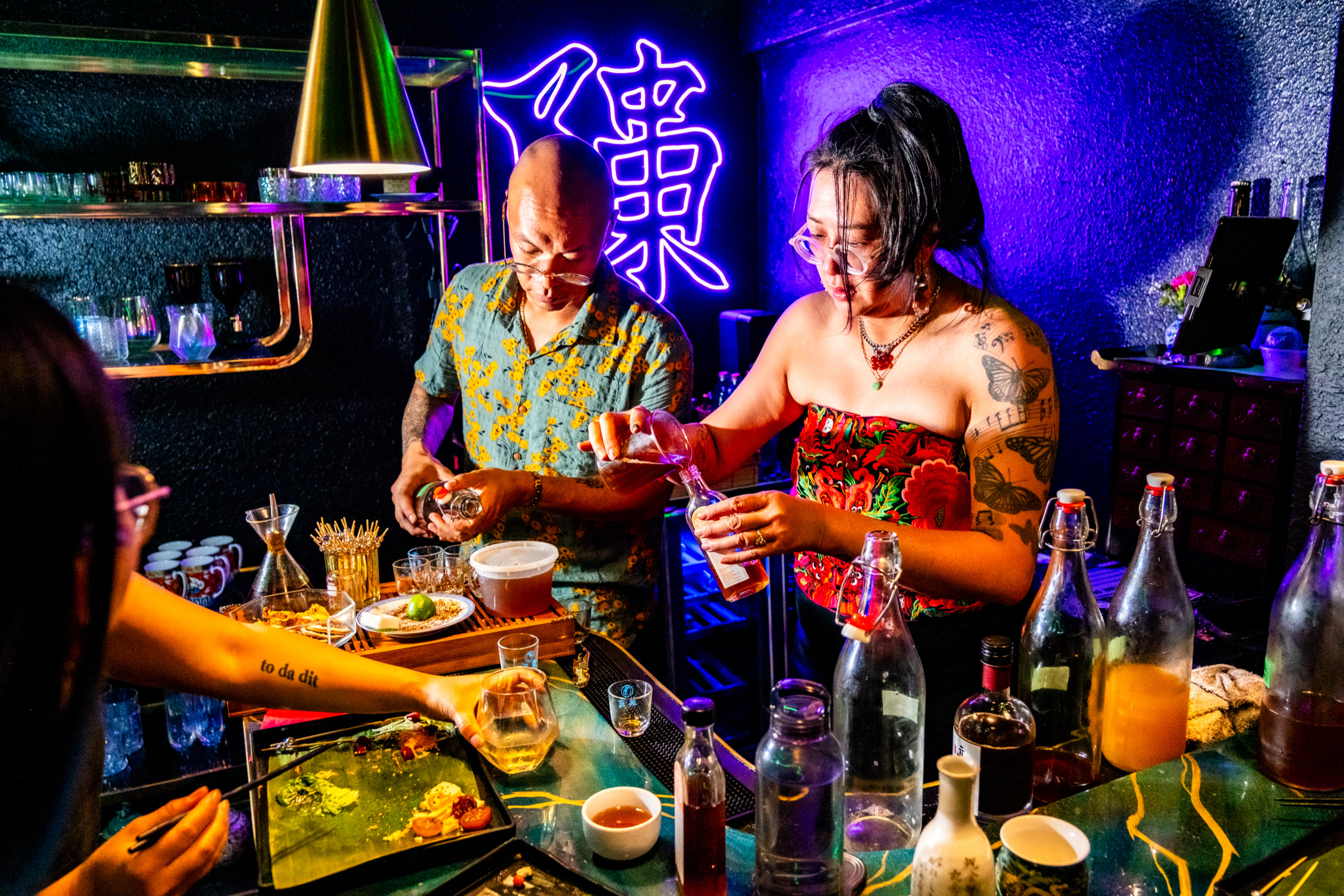 The image shows two people behind a bar mixing drinks, with neon lights and various bottles around them. A person in the foreground holds a drink.