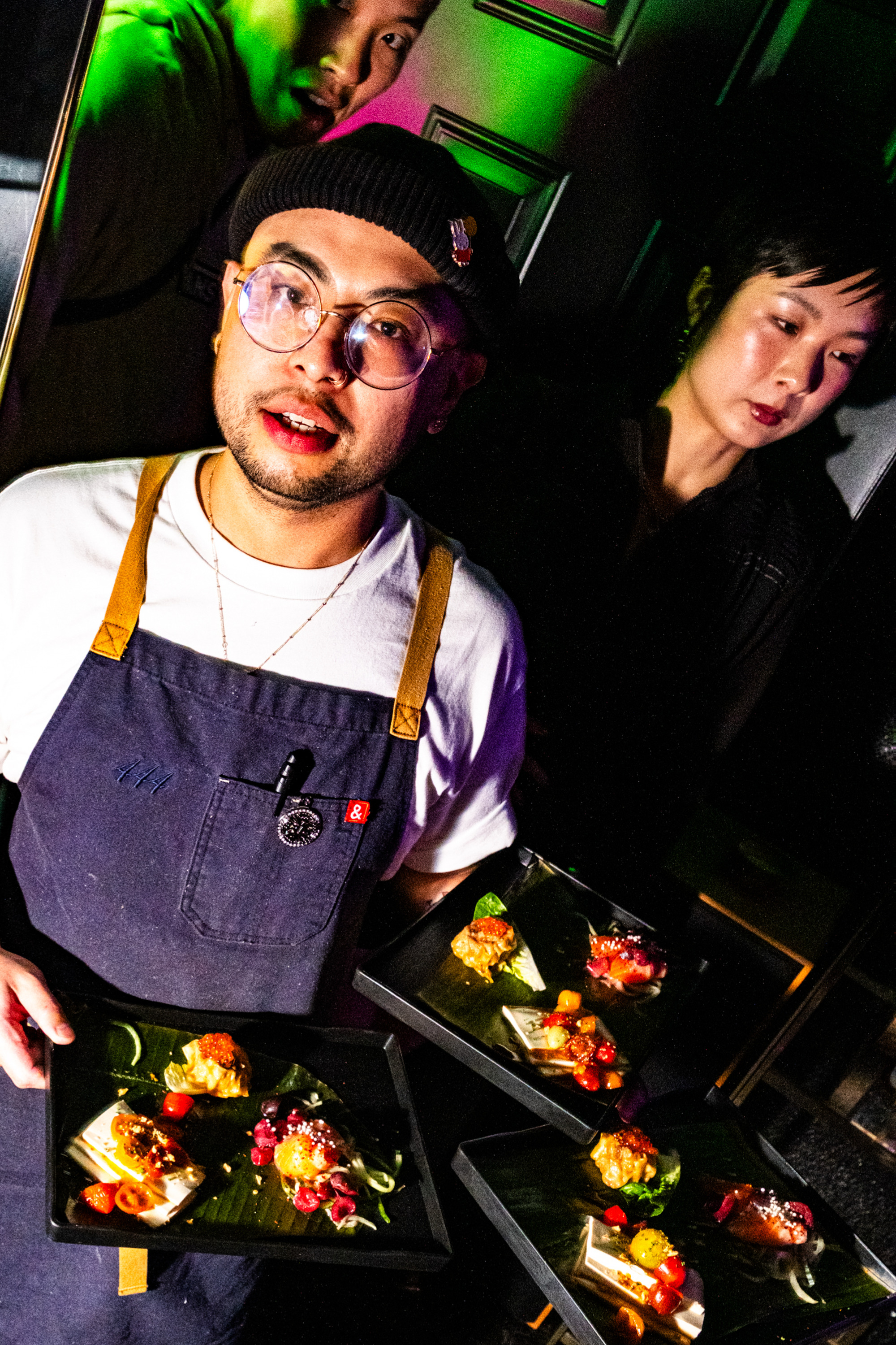 A person with glasses and a beanie holds plates of colorful, artfully presented food. Two others are in the background, with colorful lighting effects.