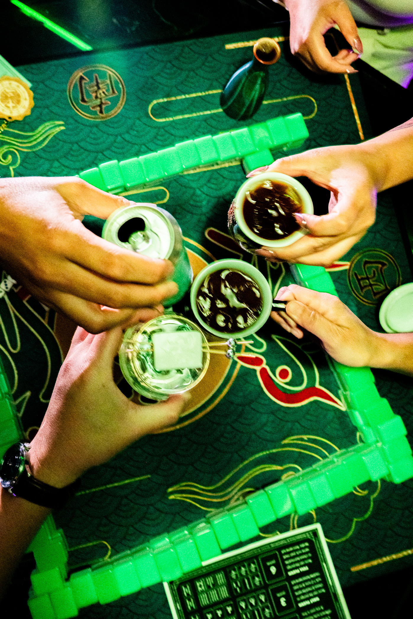 Four hands are toasting with drinks over a green mahjong table. The drinks include a can, two cups of dark liquid, and a cocktail with a straw.