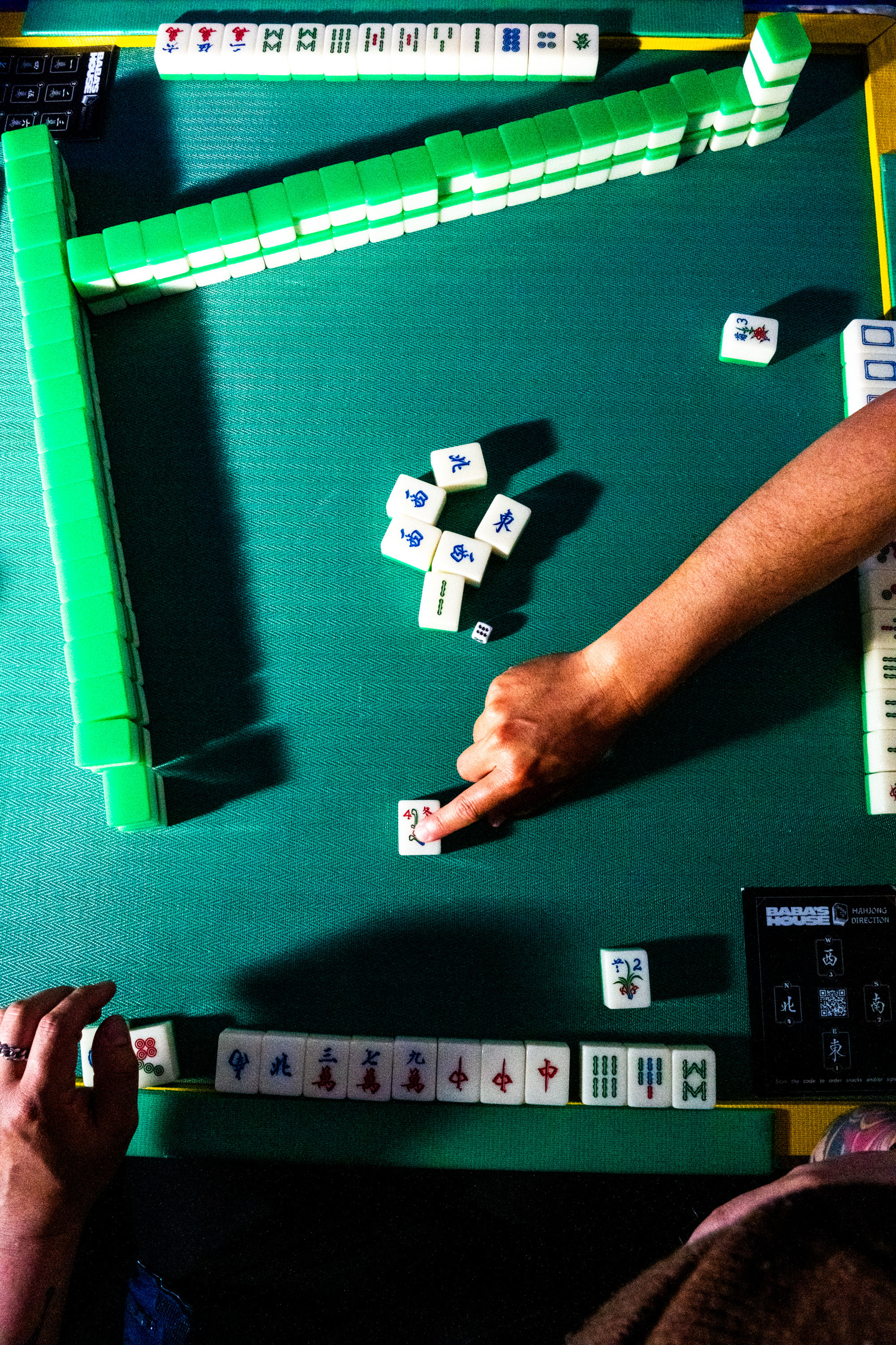 A Mahjong game is in progress on a green table. A hand points to a tile, while other tiles are arranged in lines. The game appears to be partway through.