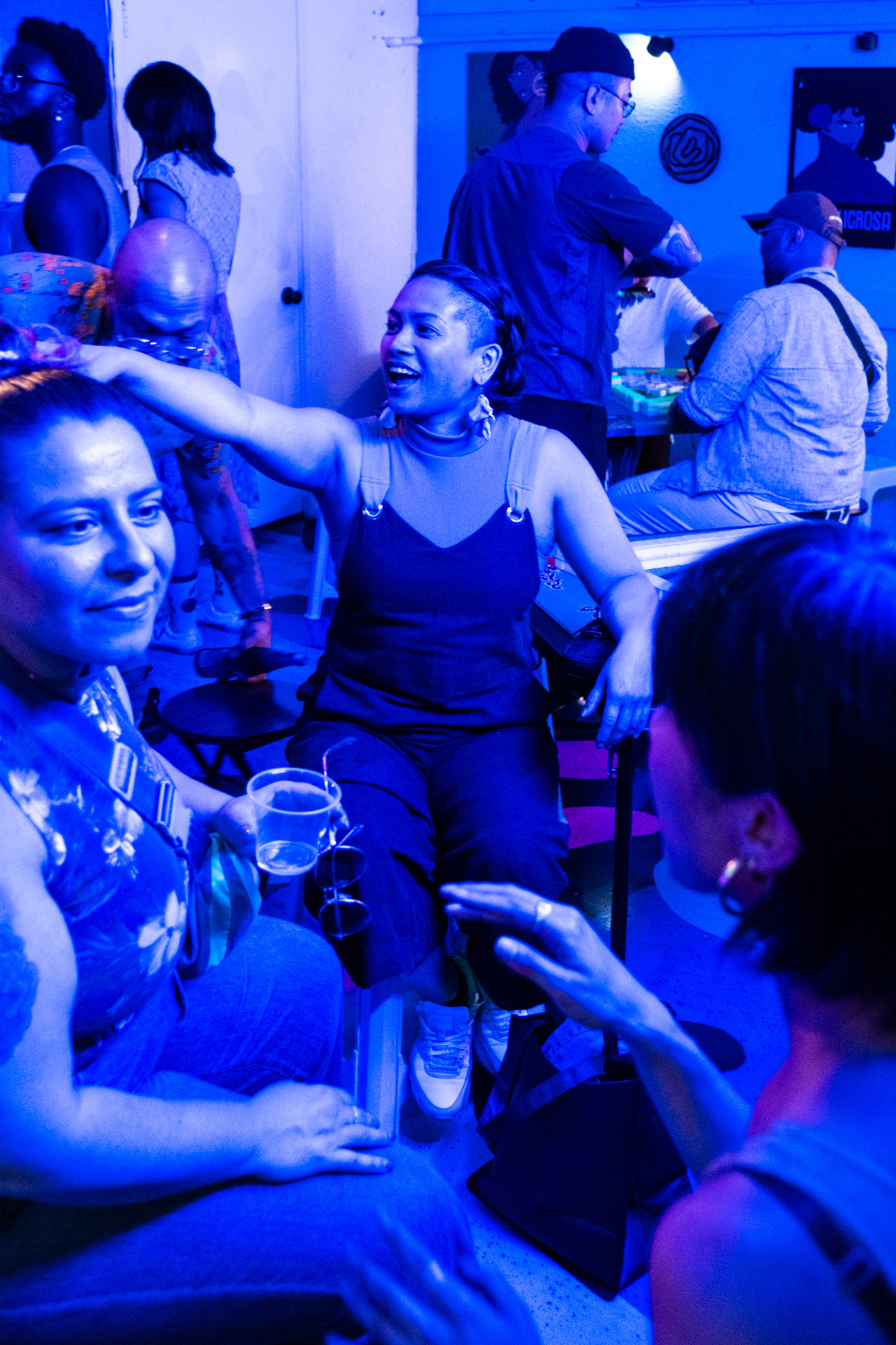 People are in a dimly lit room with blue lighting, engaging in conversation. One person is drinking from a cup, and others are interacting energetically.