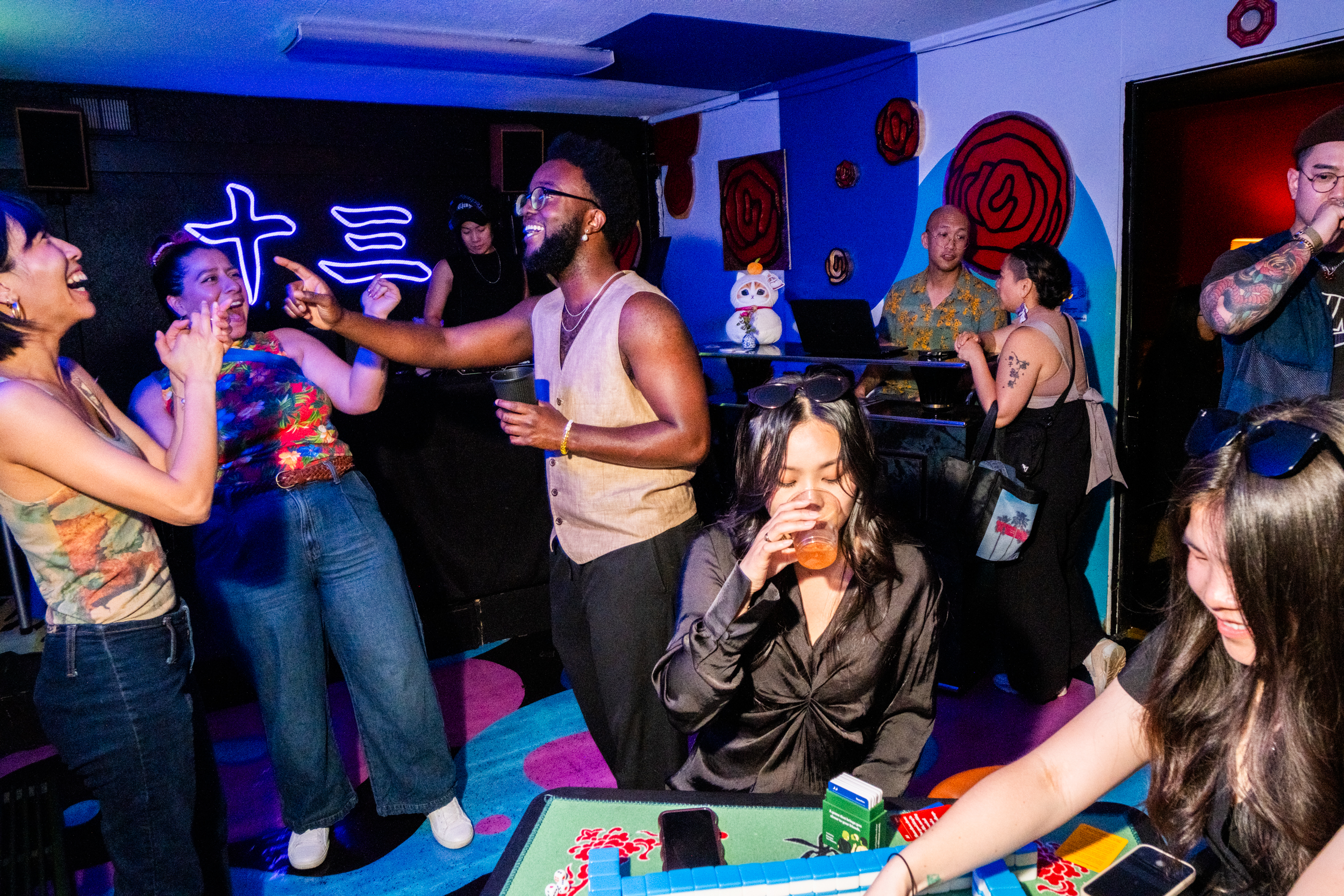 A lively party scene with people laughing and socializing. There's a DJ in the background, neon lights on the wall, and several people enjoying drinks around a table.